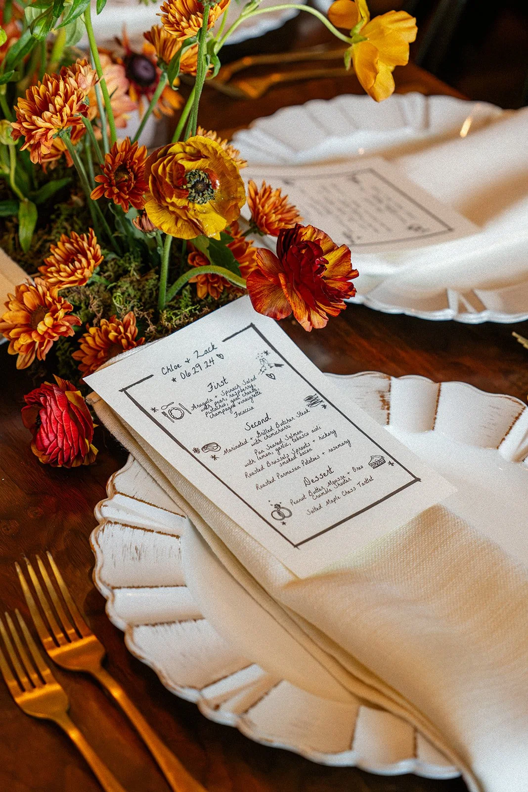 A floral centerpiece with orange and yellow flowers, a printed menu on a white napkin, and gold utensils on a wooden table.