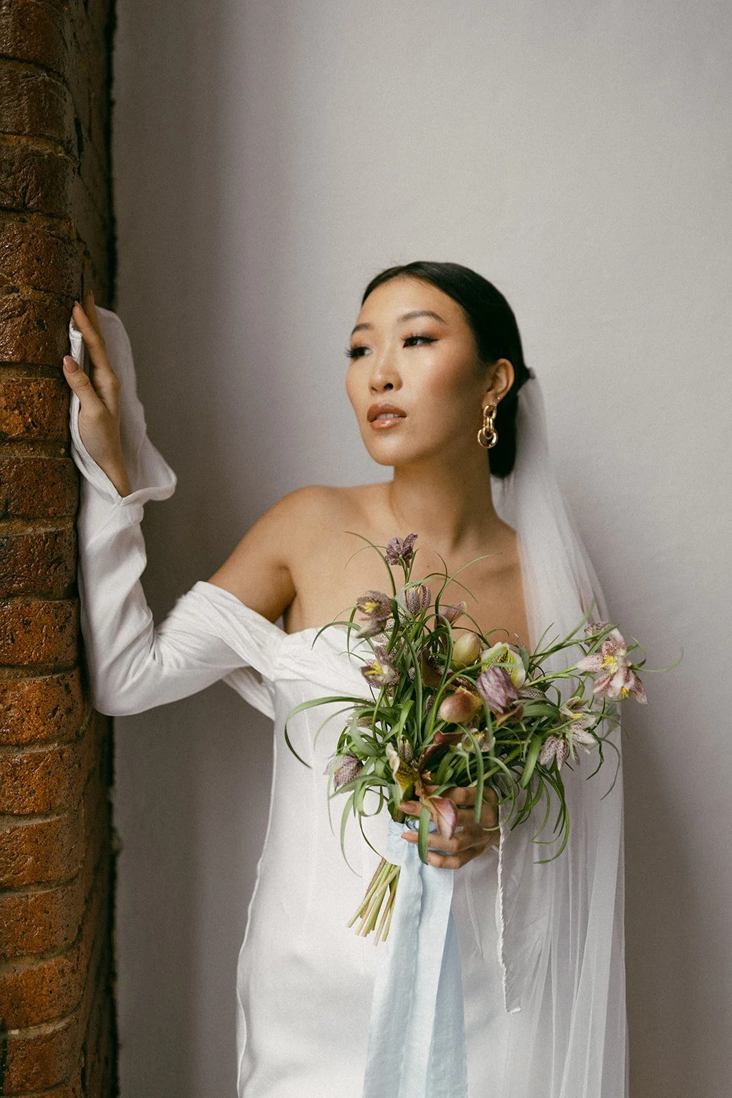 A woman in a white dress holding a bouquet of flowers, standing against a brick wall and a plain white background, with her hand on the brick wall and looking away.