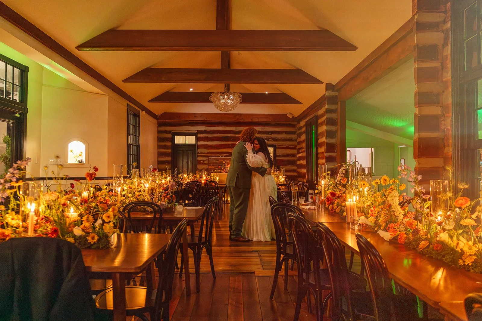 A bride and groom dance in a warmly lit reception hall decorated with flowers and candles, with rustic wood walls and a wooden ceiling.