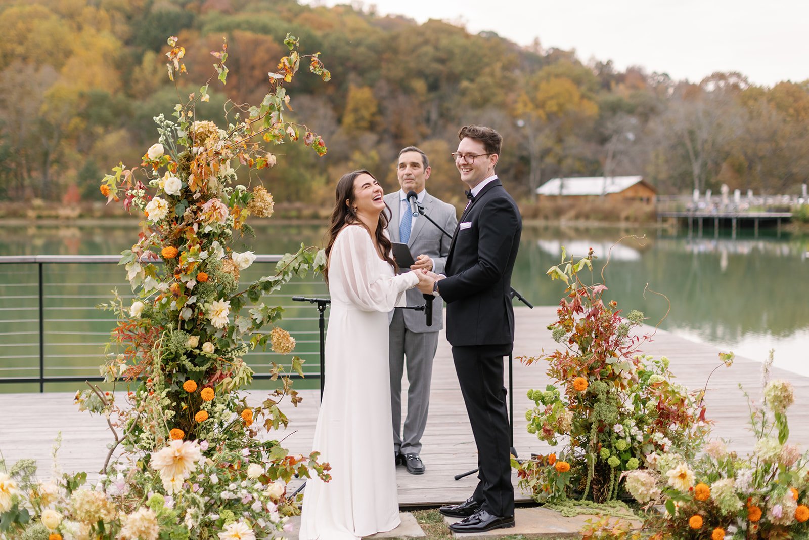 A wedding ceremony outdoors beside a lake, with a bride and groom holding hands and smiling at each other, surrounded by a floral arch and arrangements, officiant standing behind them.