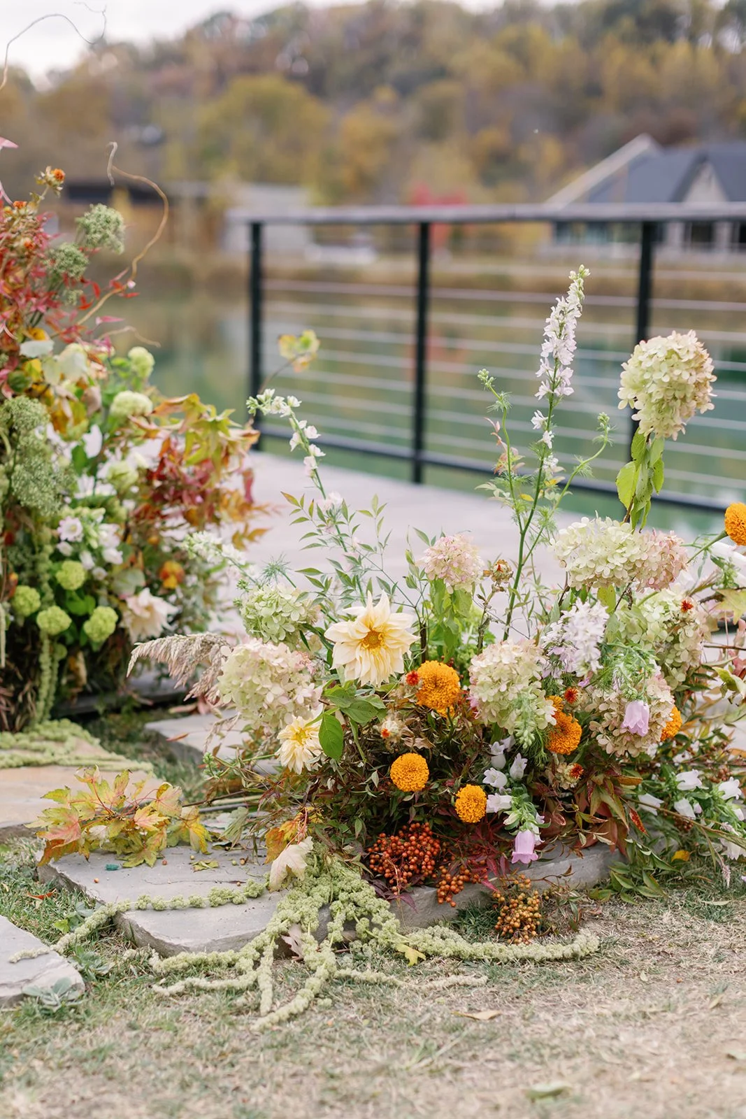 A floral arrangement with white, cream, and orange flowers near a metal railing overlooking a body of water with trees in fall foliage.