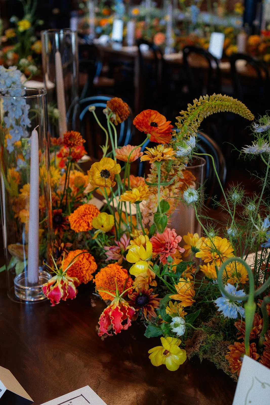 Colorful floral arrangement on a table with orange, yellow, pink, and blue flowers, set against a background of chairs and additional flowers.