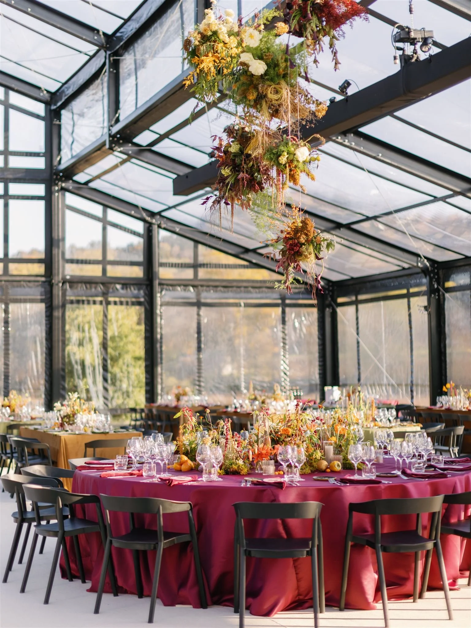 Elegant event setup inside a glass-walled venue with long tables covered in vibrant tablecloths, adorned with floral centerpieces, glassware, and plates, featuring a hanging floral arrangement above.