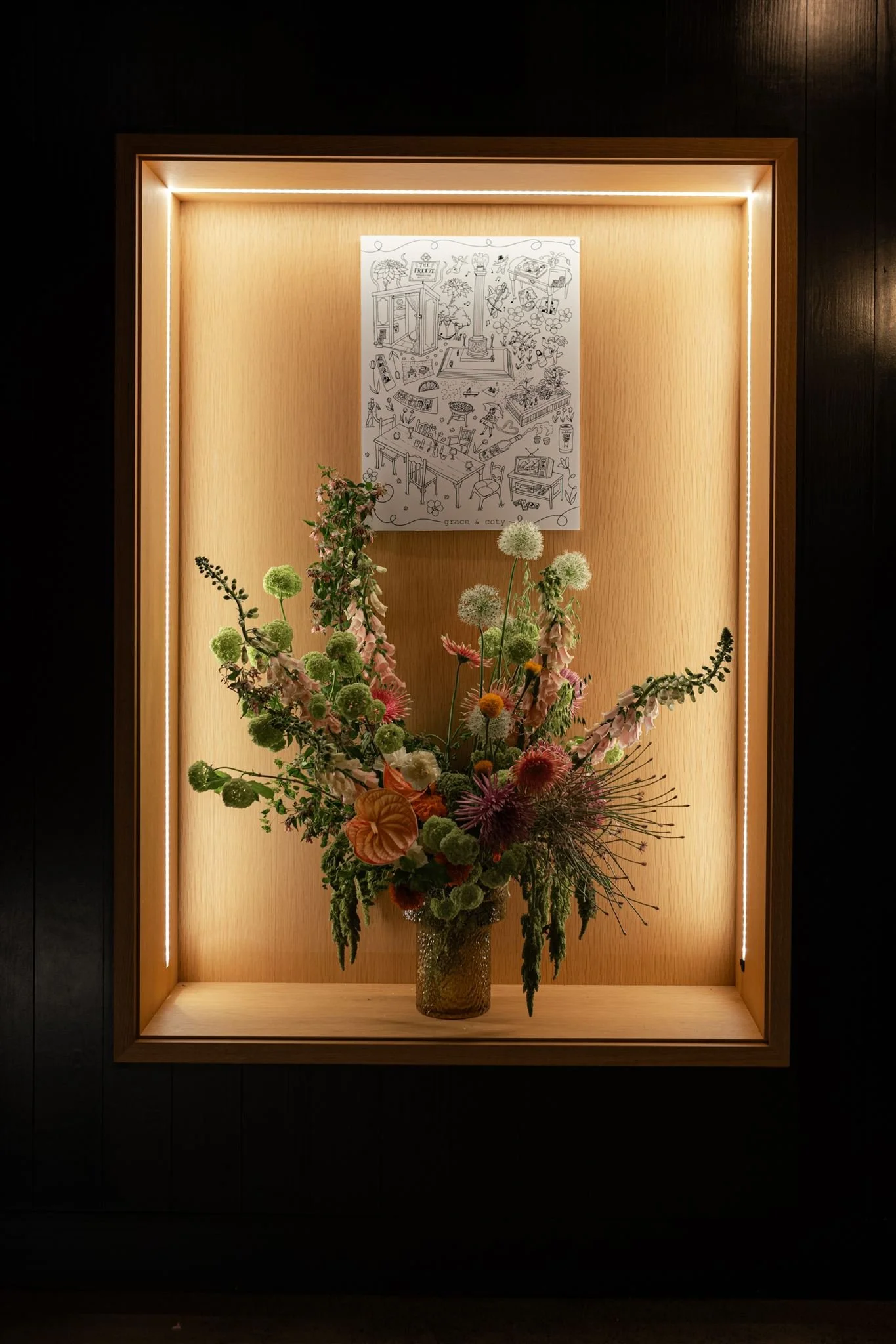 A framed floral arrangement in a glass vase displayed on a wooden shelf, with a sketch drawing on white paper above, inside a wooden display case with LED lighting.