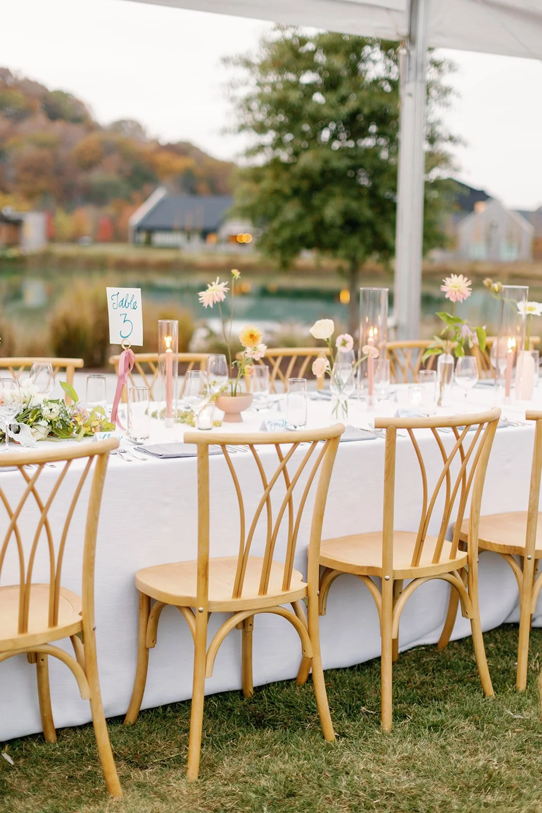 A long outdoor wedding reception table decorated with flowers and candles, set in a scenic outdoor area with trees and a body of water in the background.