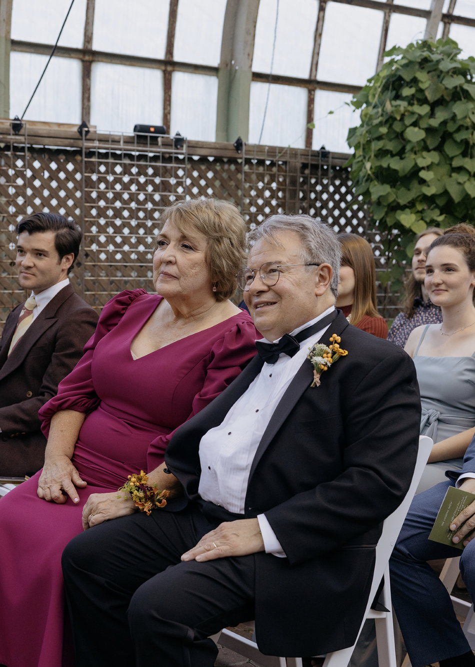 Elderly man and woman sitting in an indoor wedding ceremony, both with flower boutonnieres, guests in formal attire, wooden lattice and greenery in the background.