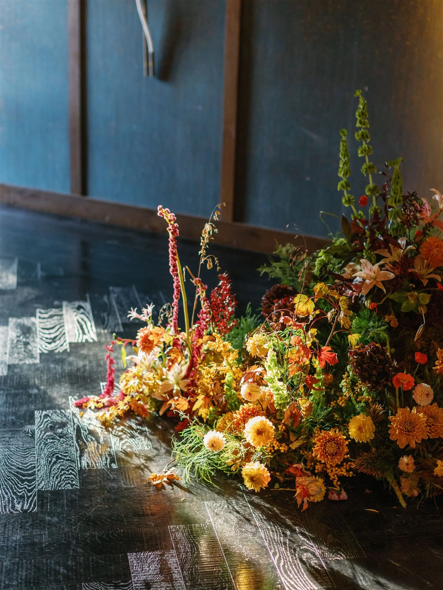 A bouquet of yellow and orange flowers with green leaves and taller pink and green floral accents on a dark wooden table.