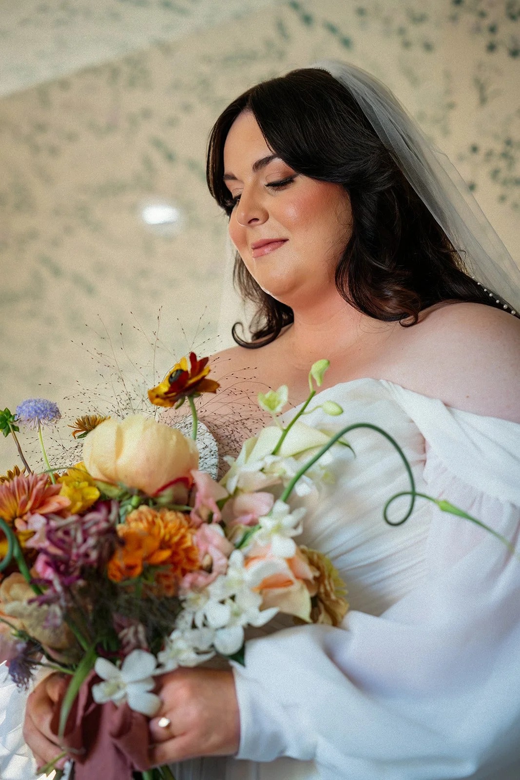 A bride with dark hair in loose curls, wearing an off-shoulder white dress and a veil, holding a colorful bouquet of flowers.