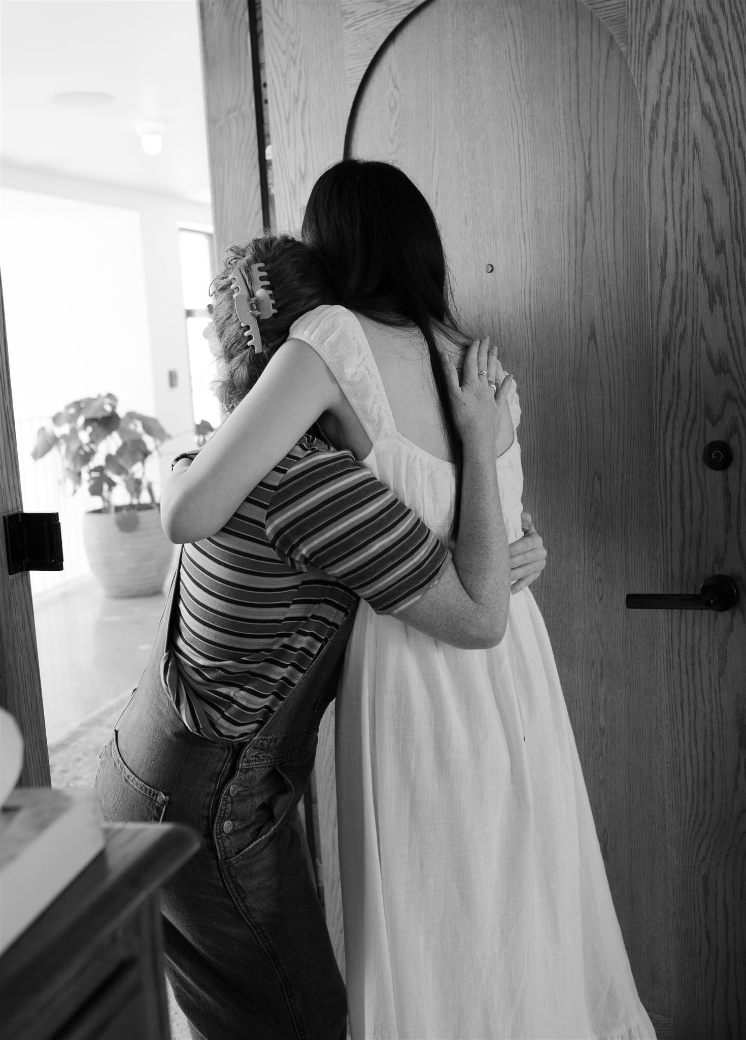 A woman in a white dress hugging a person with a striped shirt and hair rollers, inside a room near a wooden door.