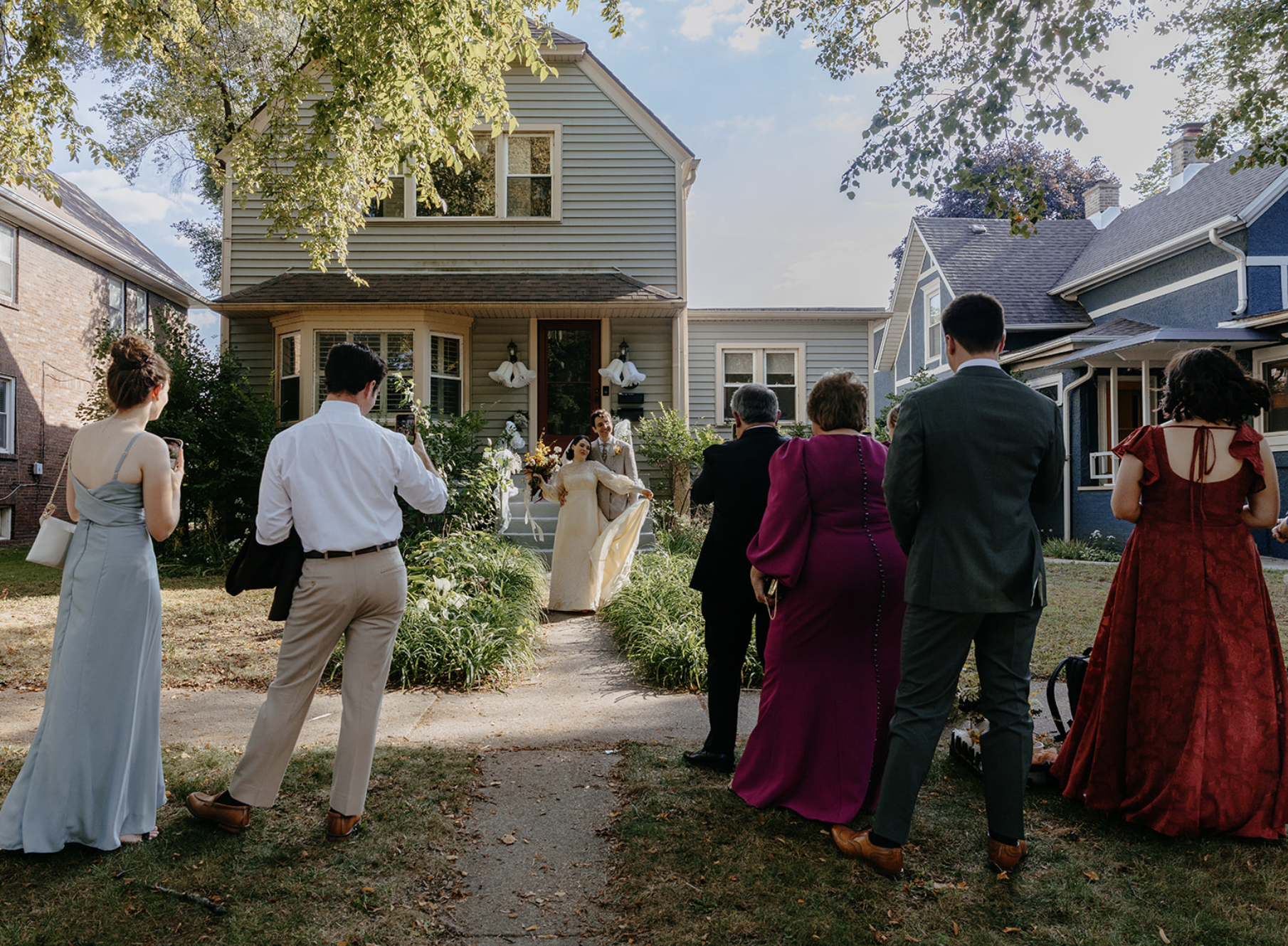 People attending a wedding outside a house, with the bride and groom standing on the front steps, while guests gather on the lawn taking photos in daylight.
