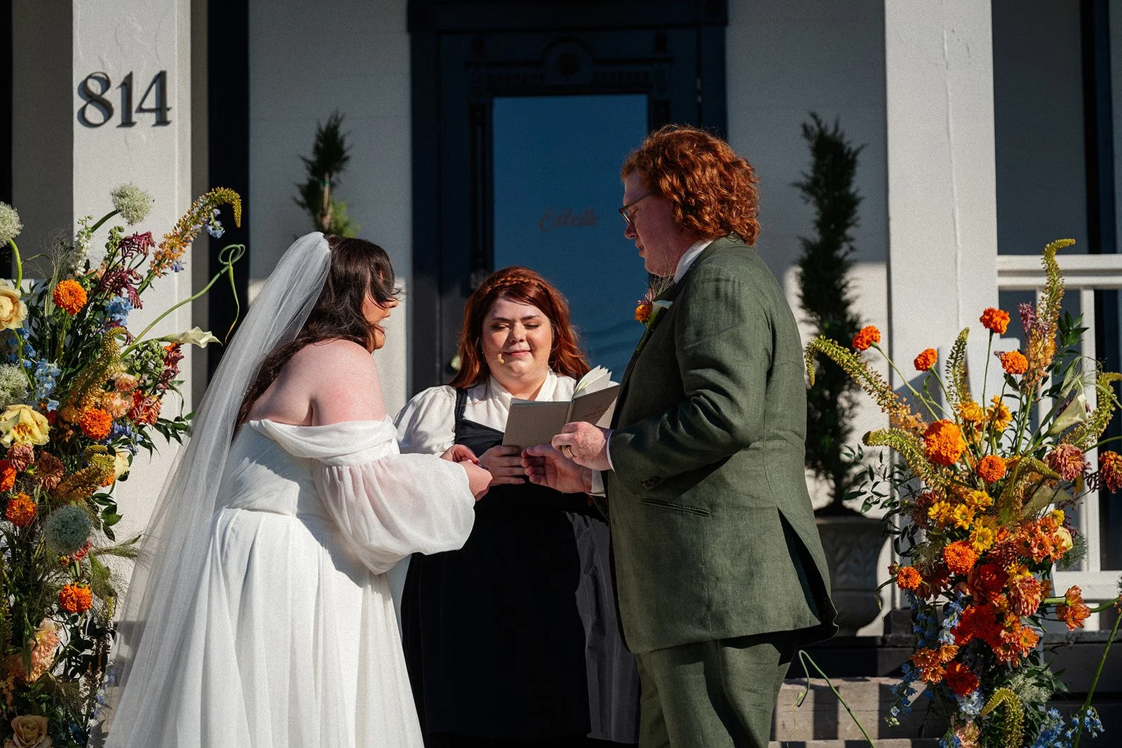A bride and groom exchanging vows during a wedding ceremony outside a house, with a woman officiant holding a booklet, surrounded by colorful flowers.