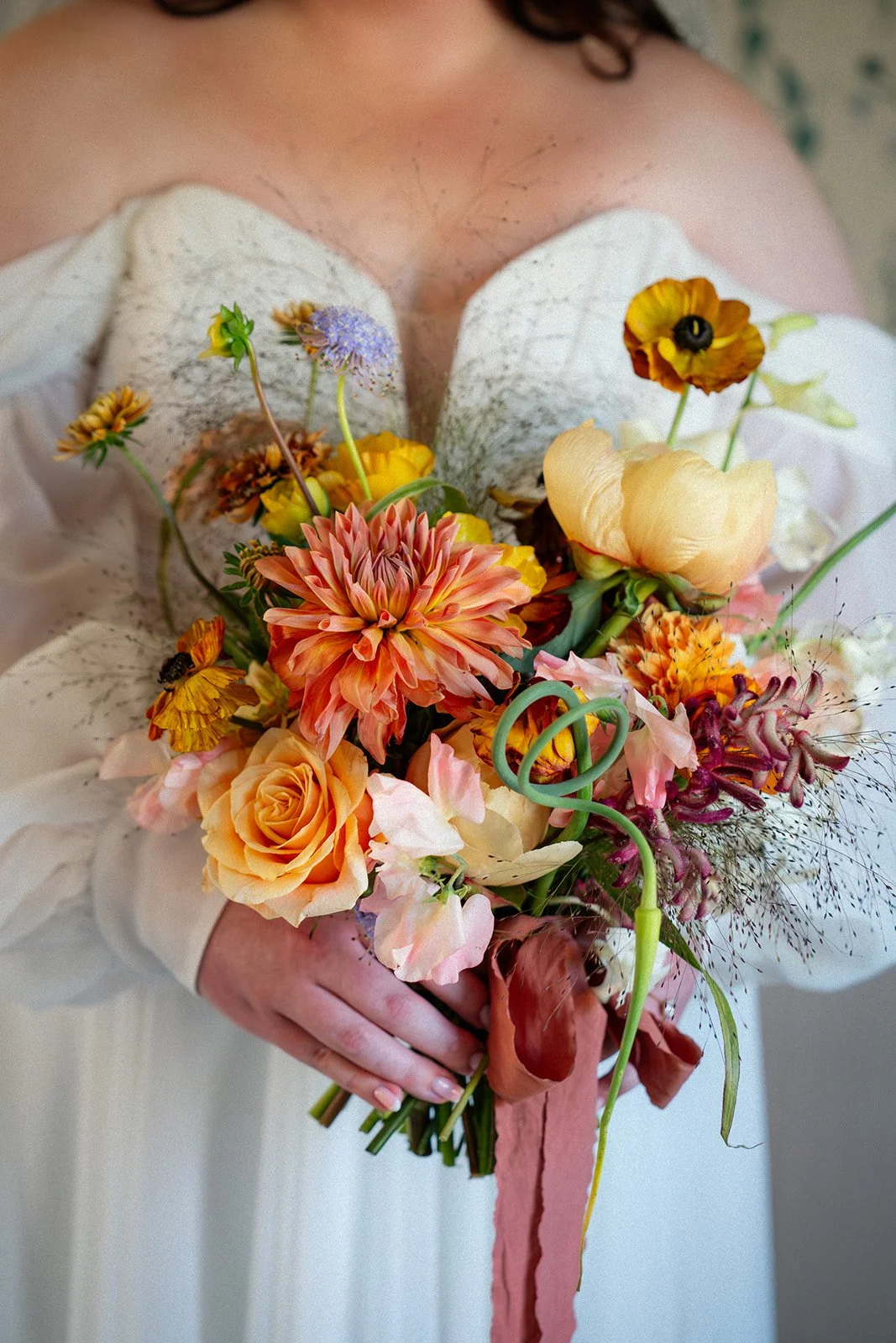 Close-up of a person holding a colorful bouquet of flowers, including roses, dahlias, poppies, and other mixed blooms, wrapped with a pink ribbon.