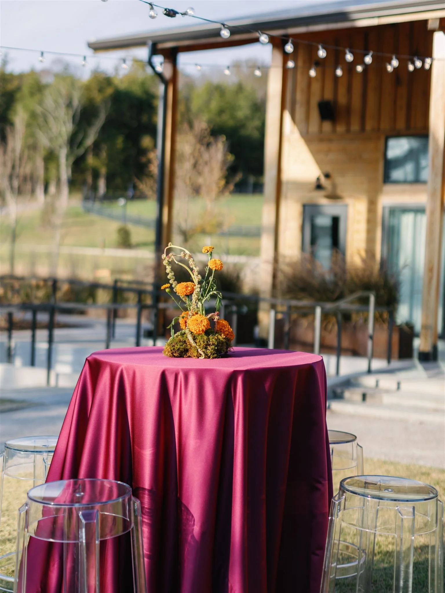 A round table with a pink satin tablecloth and a floral centerpiece with orange and yellow flowers outdoors, with transparent acrylic chairs around it, under string lights, near a wooden house with a chimney, during daytime.