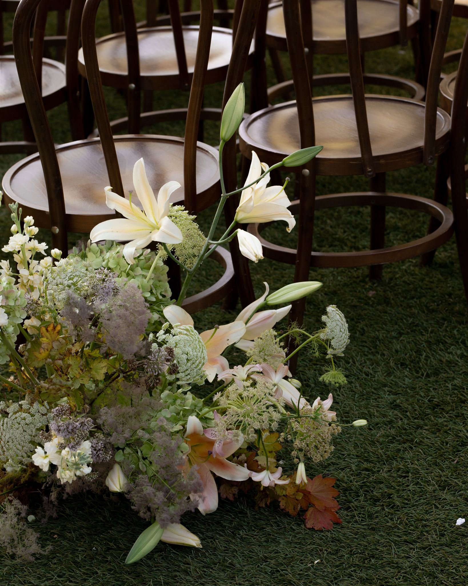 A floral arrangement of white lilies and other mixed flowers on green grass with wooden chairs in the background.