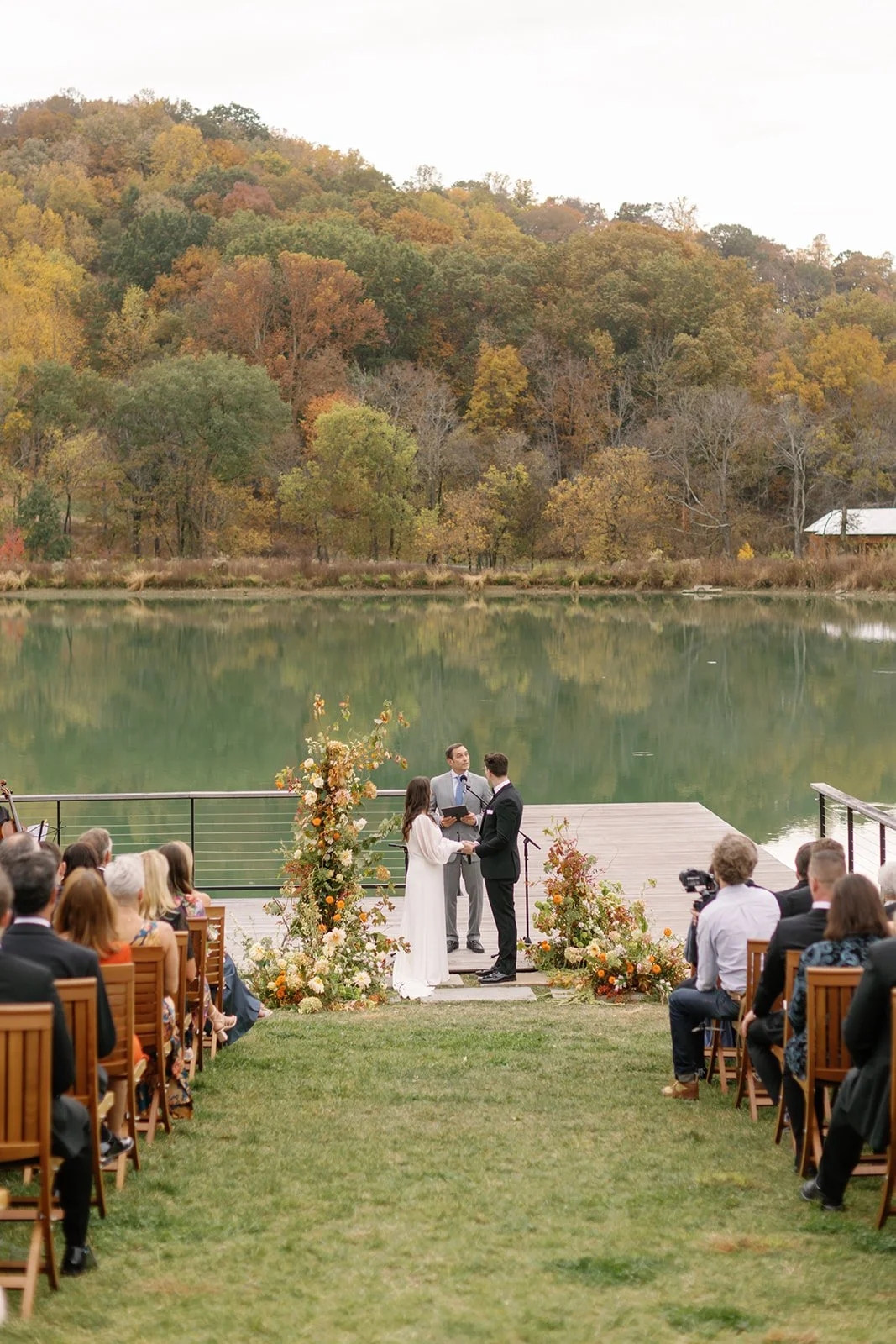 A wedding ceremony taking place outdoors by a lake with autumn foliage in the background. The bride and groom are holding hands and exchanging vows in front of an officiant, surrounded by floral arrangements. Guests are seated on both sides of a gras