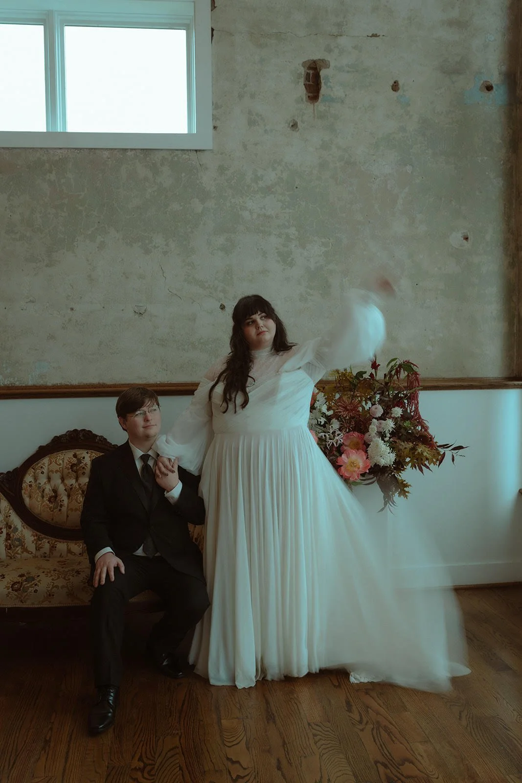 A woman in a white gown and a man in a black suit sitting on a vintage sofa in a room with a large window. The woman is holding the man's hand and raising her arm, while the man is looking at her. There is a colorful flower arrangement behind them, i
