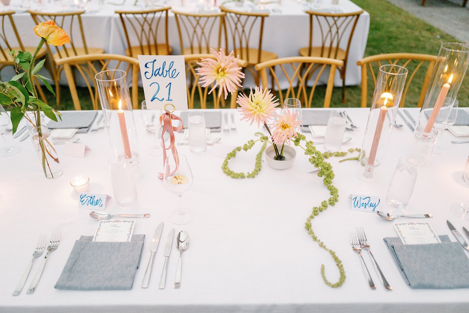 An elegantly decorated outdoor wedding reception table with a white tablecloth, pink flowers, candles, and table number '21' sign in the center. Place settings include gray napkins, silverware, and clear glassware. There are chairs with wood backs ar