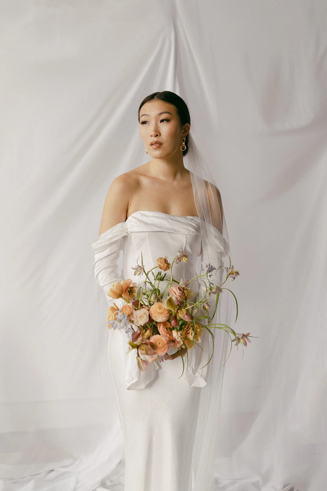 A woman in a white off-the-shoulder dress holding a bouquet of pink and purple flowers, standing against a white draped background.