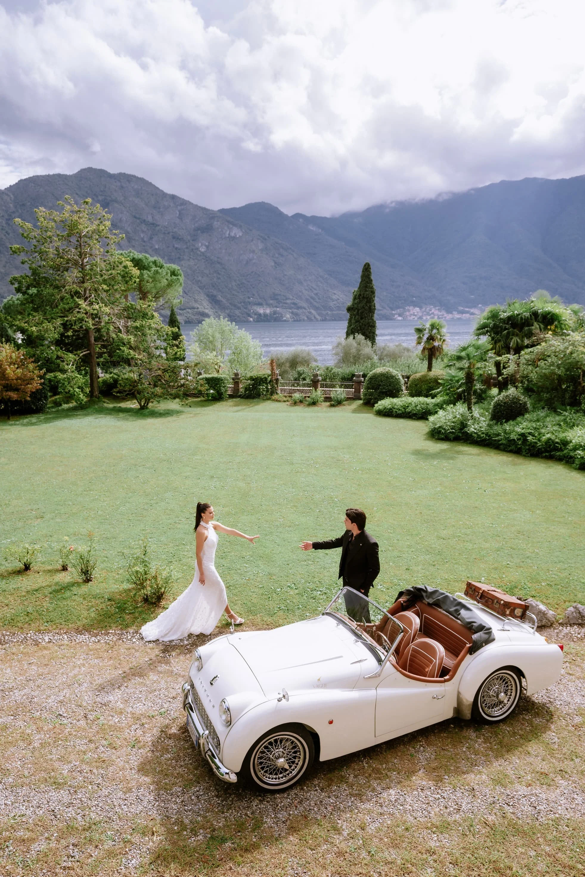 A bride and groom dancing beside a vintage white convertible car on a lush green lawn with mountains and a lake in the background.