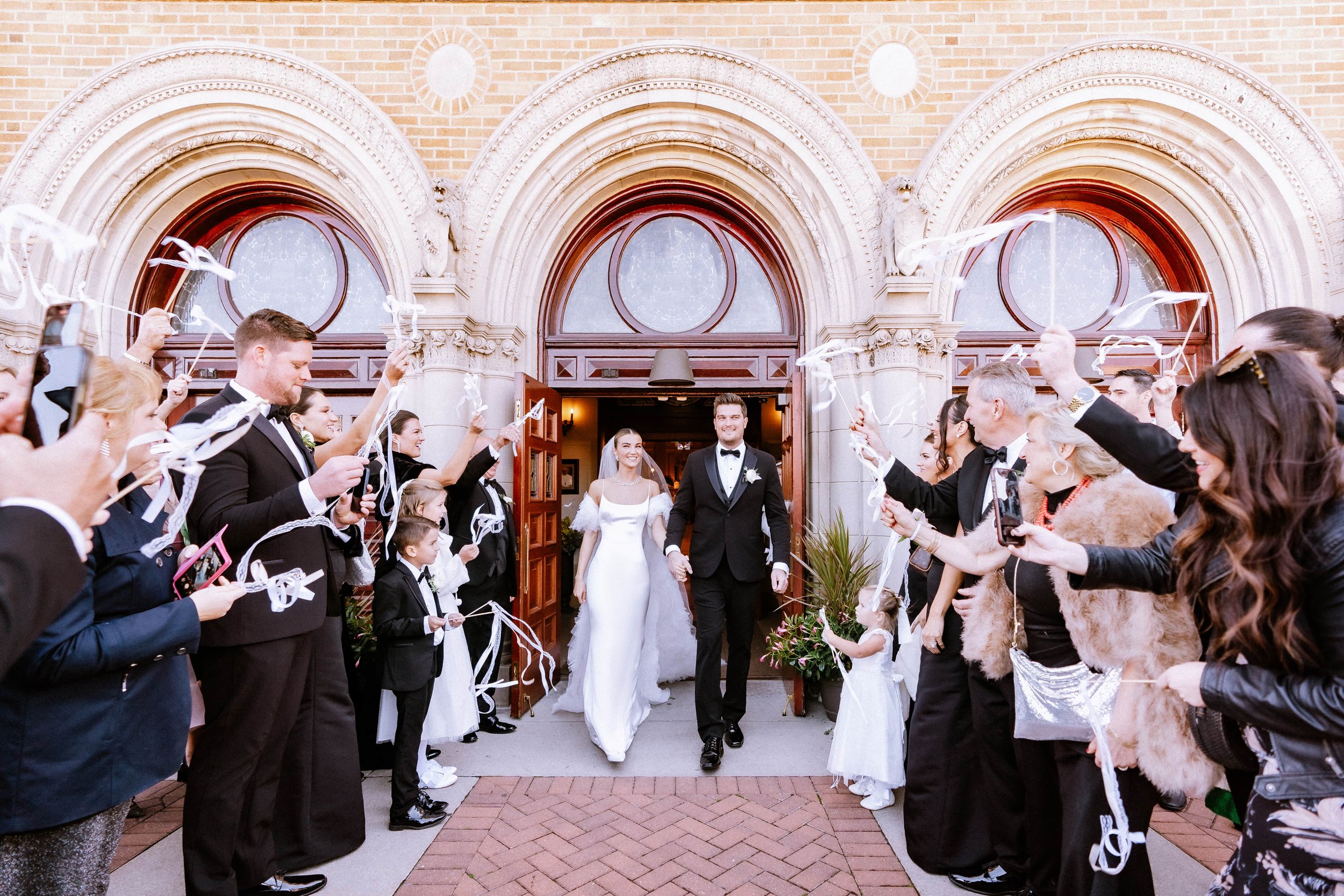 Bride and groom walking out of a church, surrounded by wedding guests celebrating with streamers and taking photos.