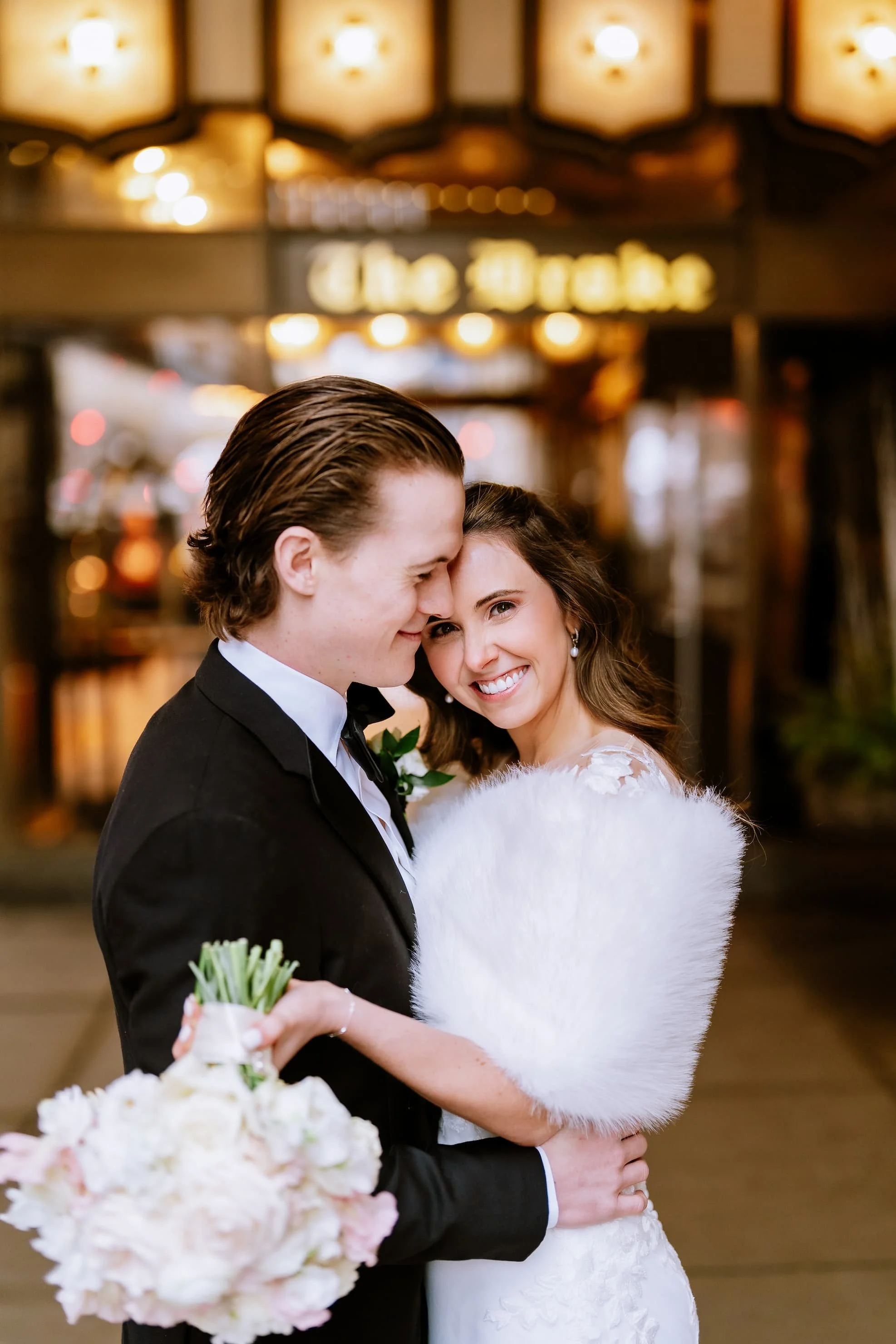A romantic wedding photo of a smiling bride and groom embracing outside a venue at night, with warm lighting and blurred background.