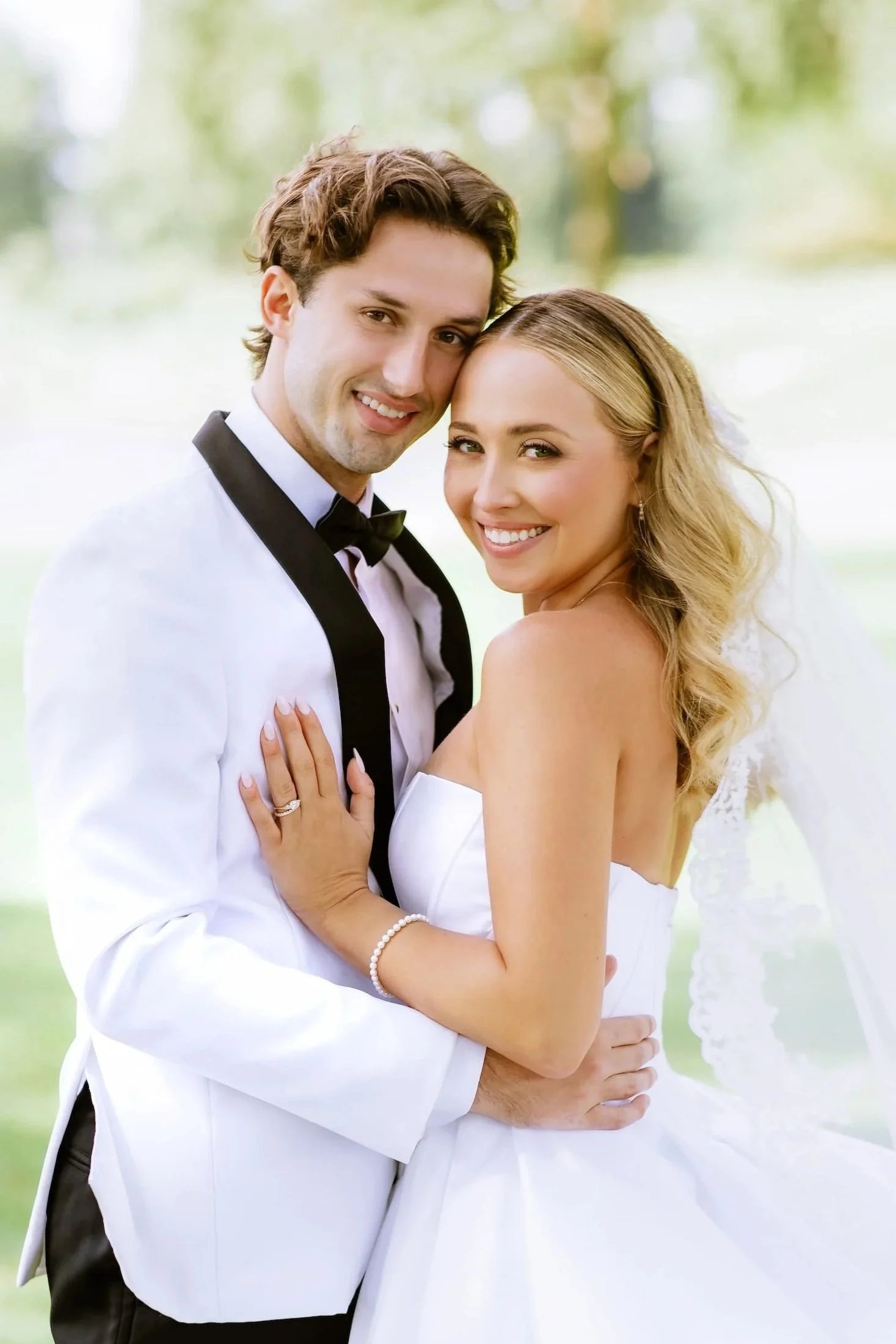 A happy bride and groom smiling, dressed in wedding attire outdoors with blurred greenery in the background.