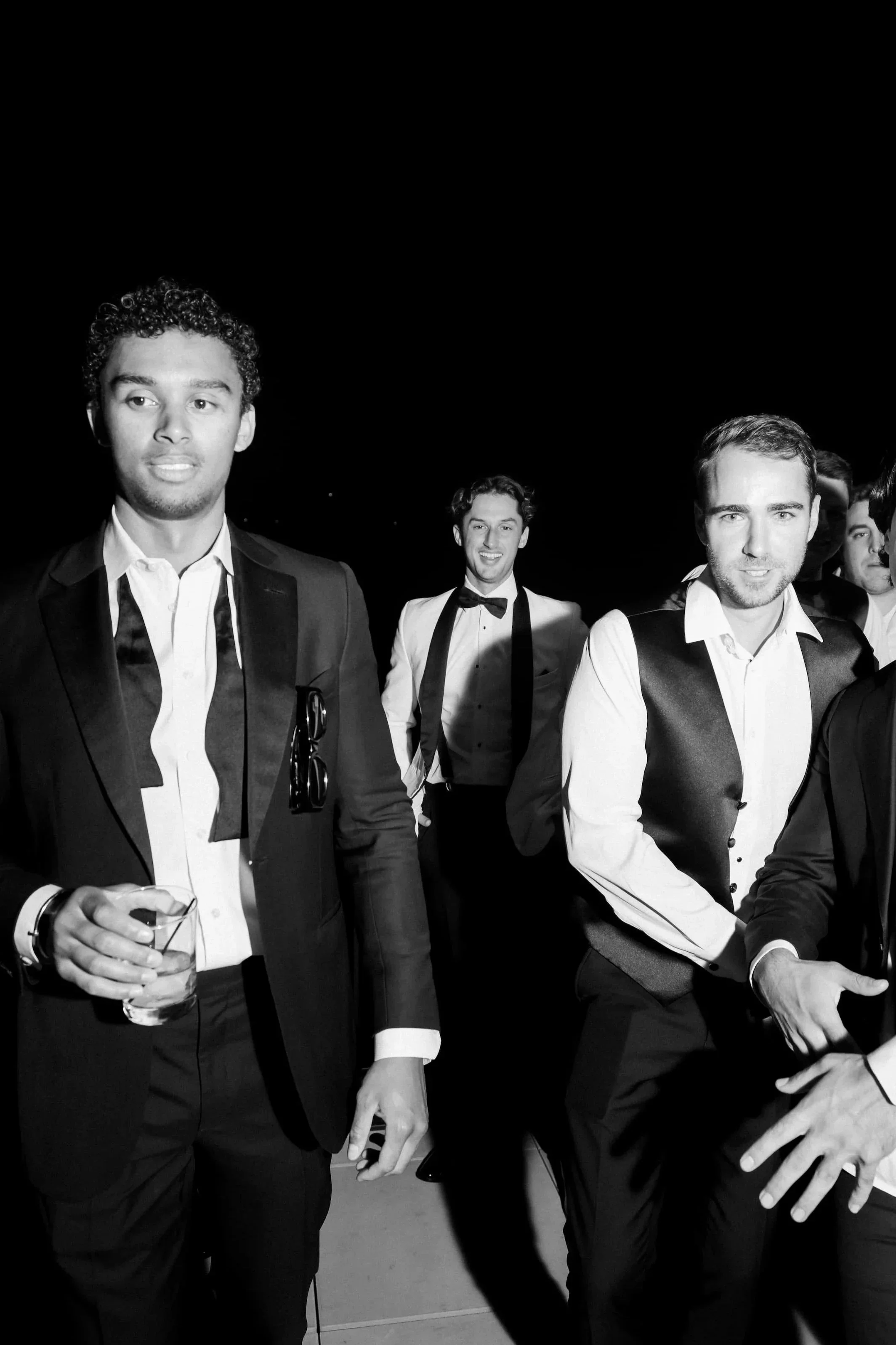 Black and white photo of a group of young men in tuxedos at a formal event, some holding drinks, smiling and enjoying themselves.