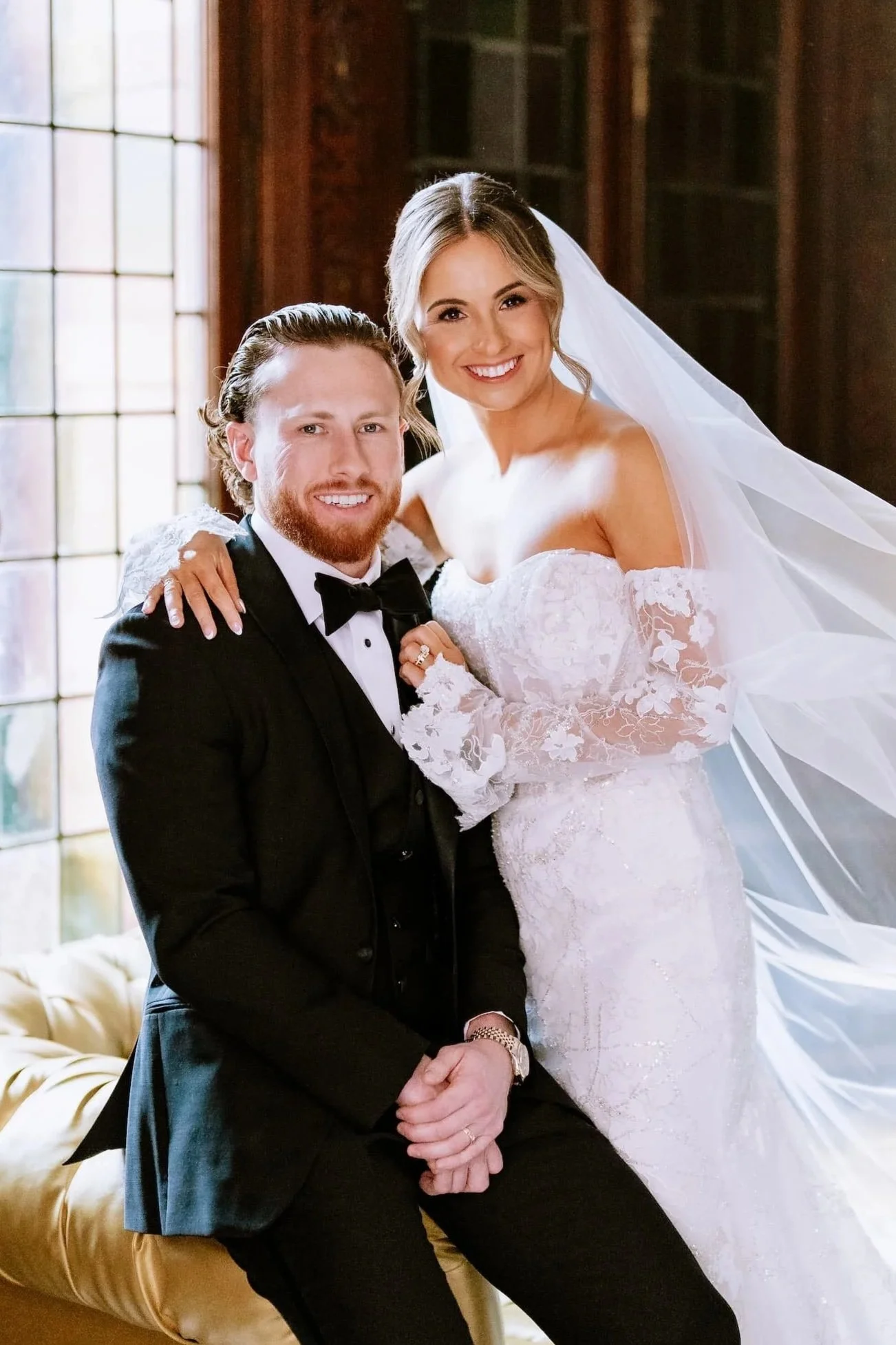 A bride in a white wedding gown with lace details and a long veil, and a groom in a black tuxedo with a bow tie, smiling and posing together indoors near a window.