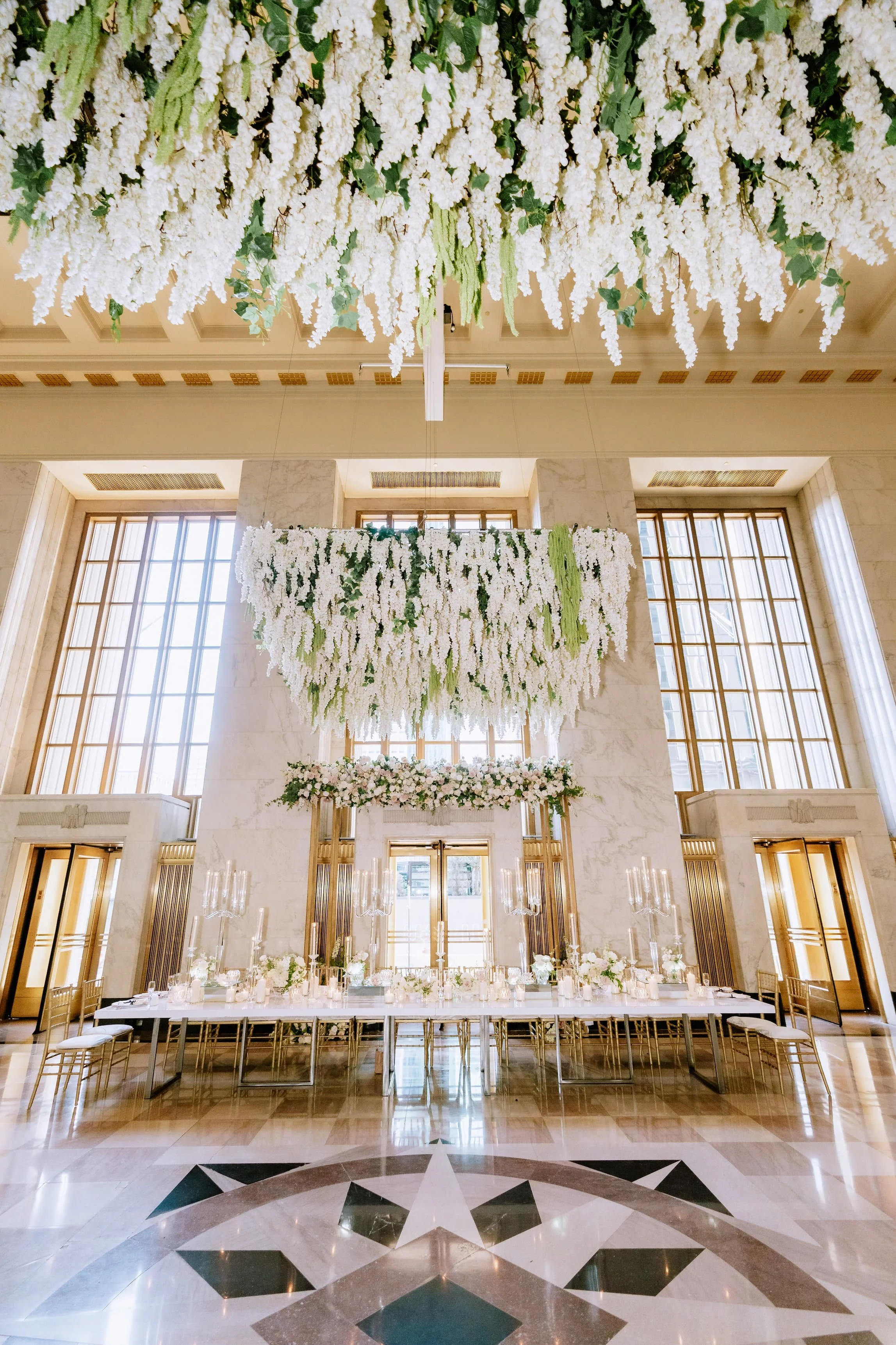 Elegant banquet hall decorated with hanging white floral arrangements, large windows with wooden frames, and a long table set for a formal event with candles and flowers.