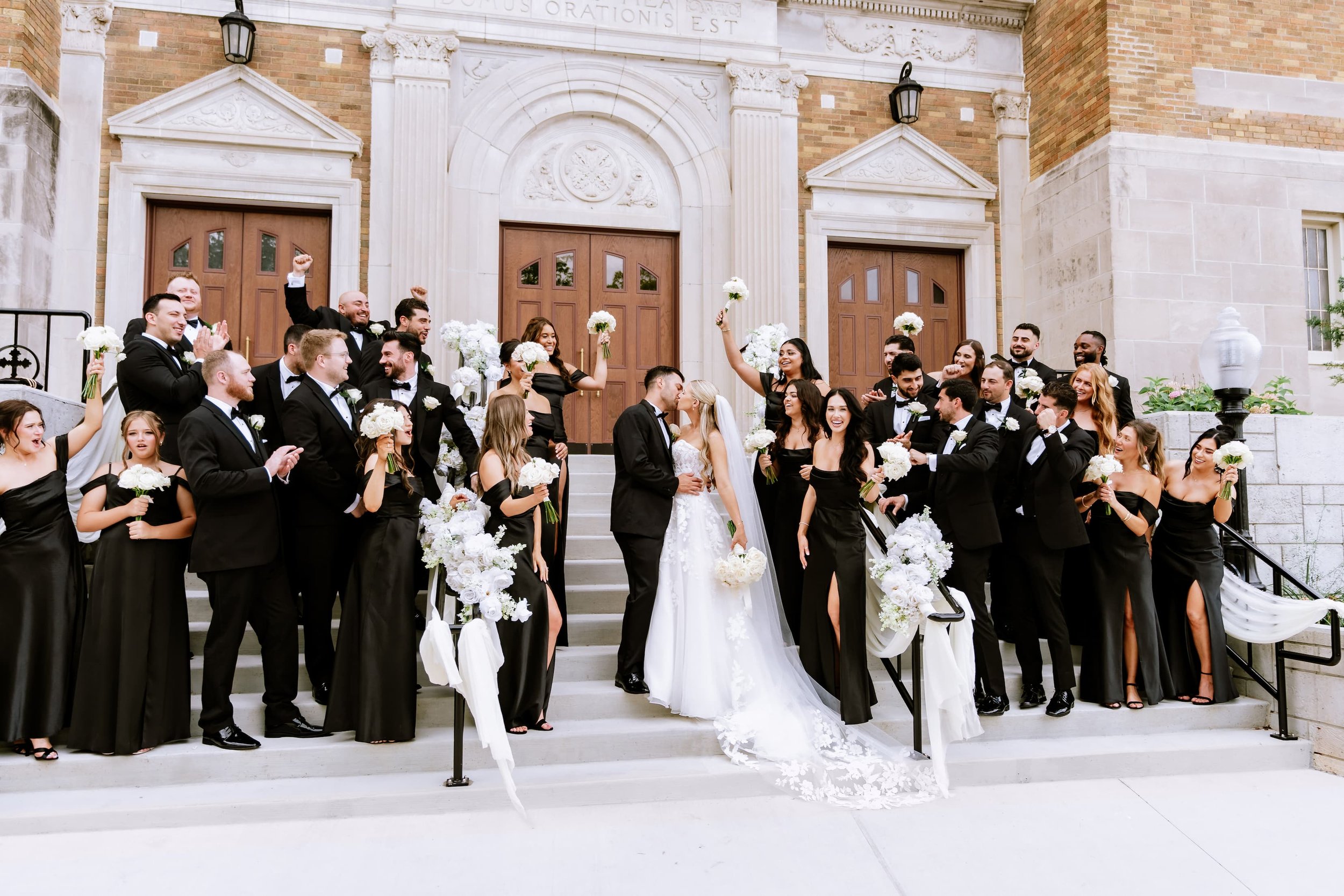 A wedding party on church steps with a bride and groom kissing, surrounded by bridesmaids and groomsmen celebrating, holding bouquets and dressed in formal black and white attire.