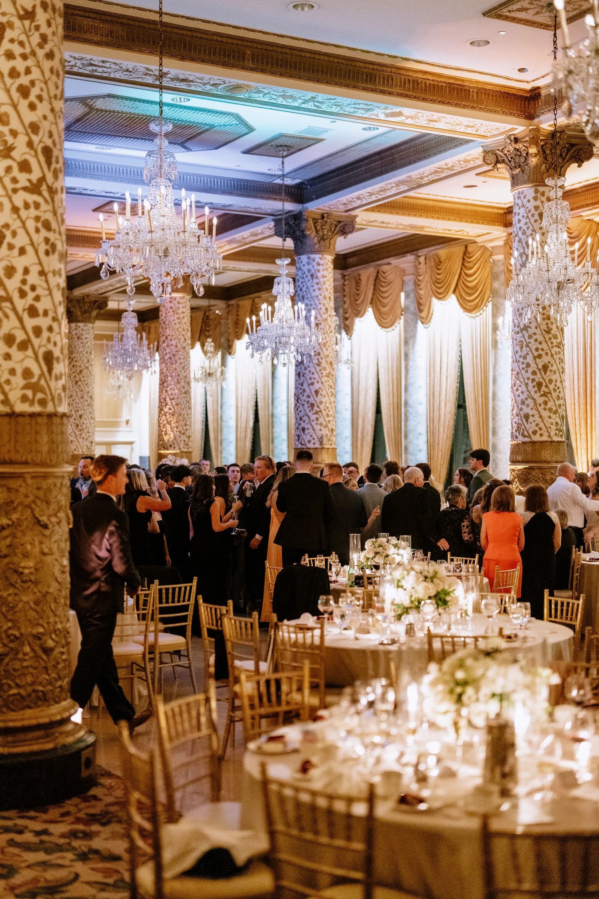 A luxurious banquet hall filled with elegantly dressed people, with chandeliers, ornate columns, golden accents, and decorated tables with white flowers and candles.