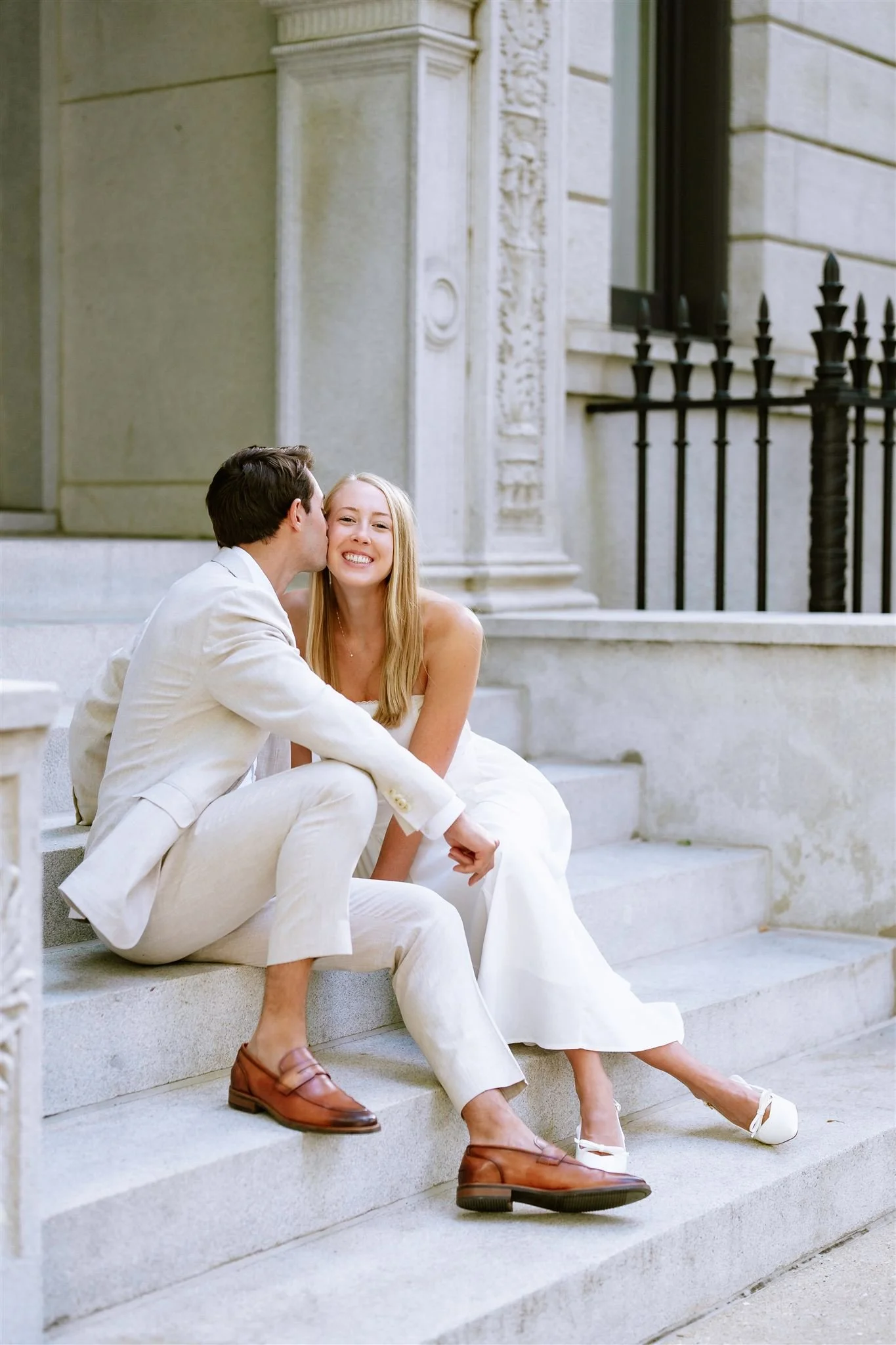 A young couple dressed in white sitting on outdoor stairs, with the man kissing the woman on the cheek. The woman is smiling, and they are near a historic stone building with ornate architectural details and black iron railing.