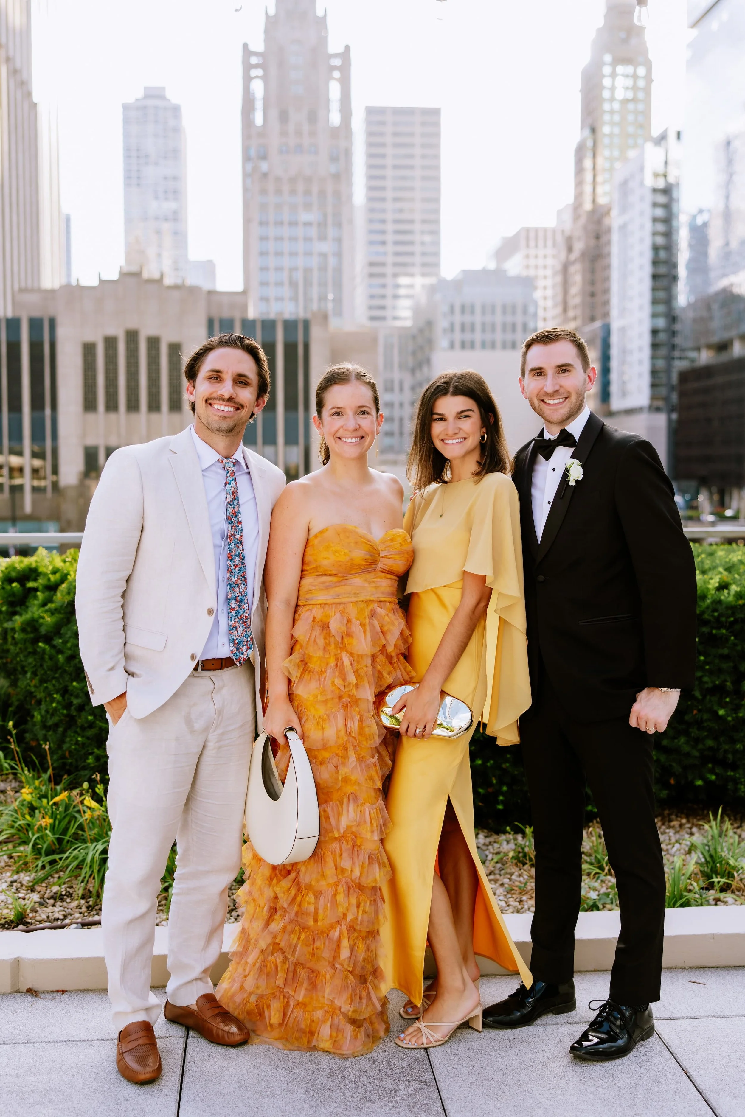 Group of four friends dressed in formal and colorful outfits, smiling outdoors in an urban setting with tall buildings in the background.
