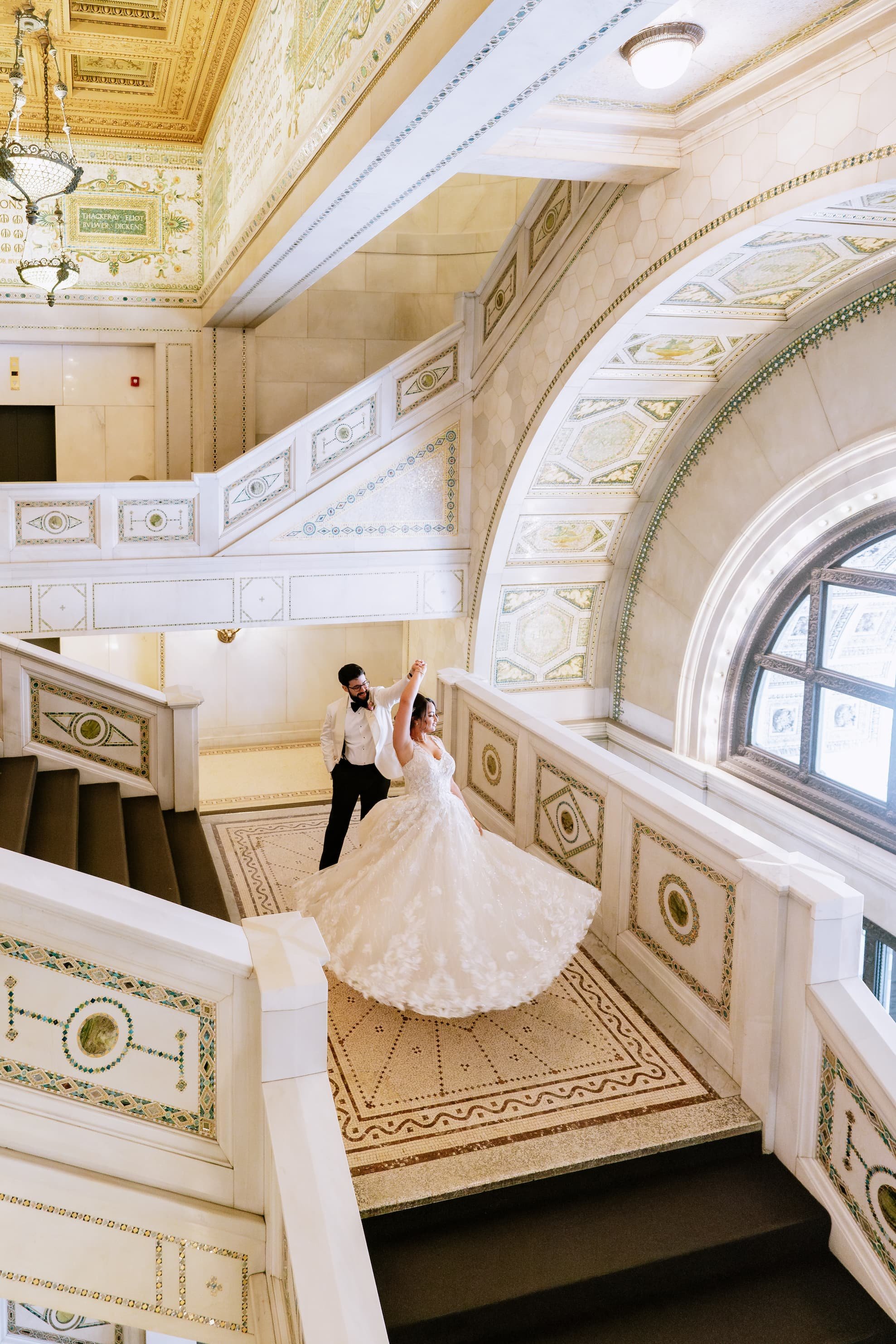 A bride and groom dancing on a grand staircase inside a historic building with ornate decorations and large arched windows.