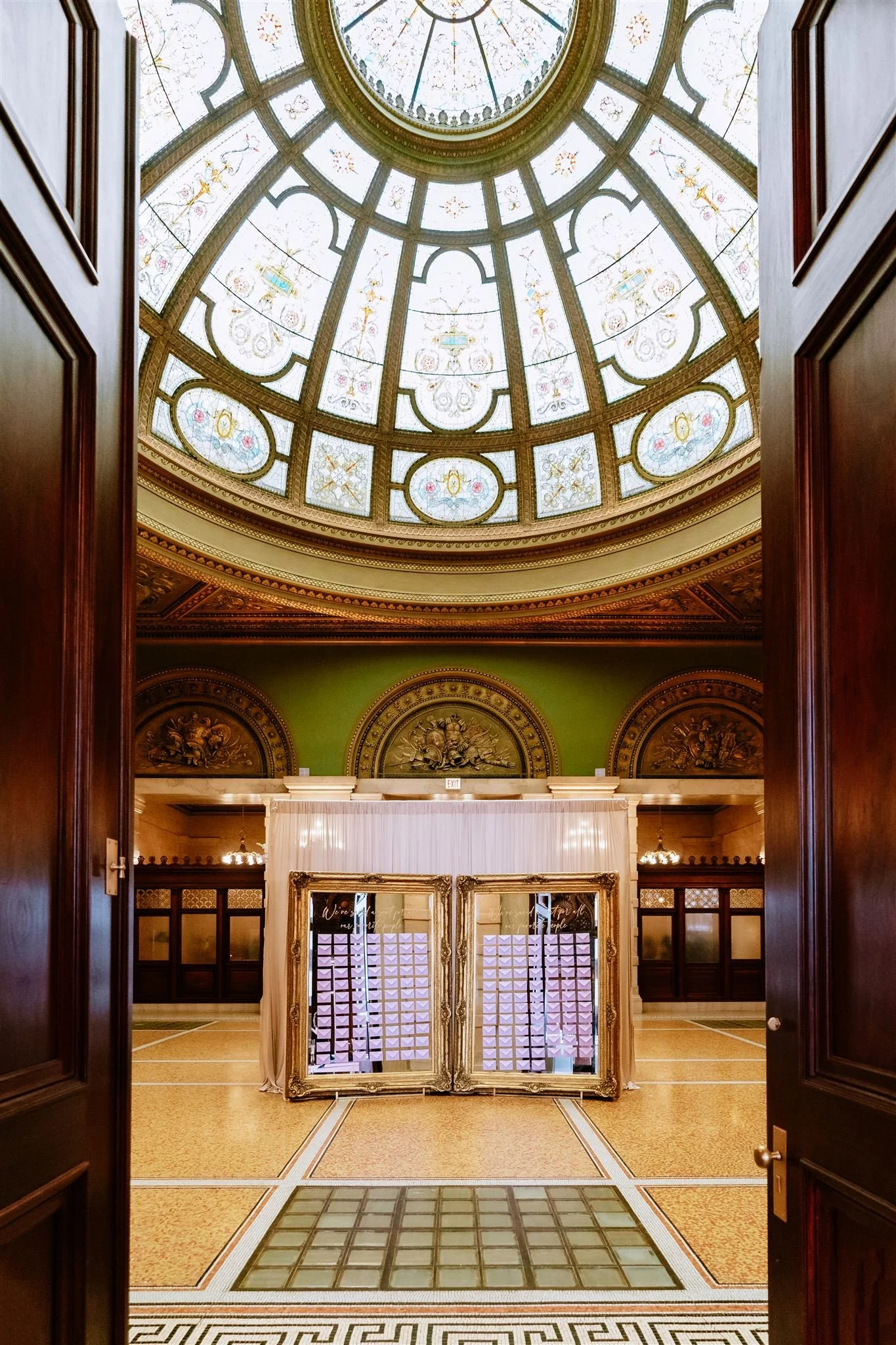 View of an elegant indoor space with a stained glass dome ceiling, ornate gold and wood details, and a mirror reception desk.
