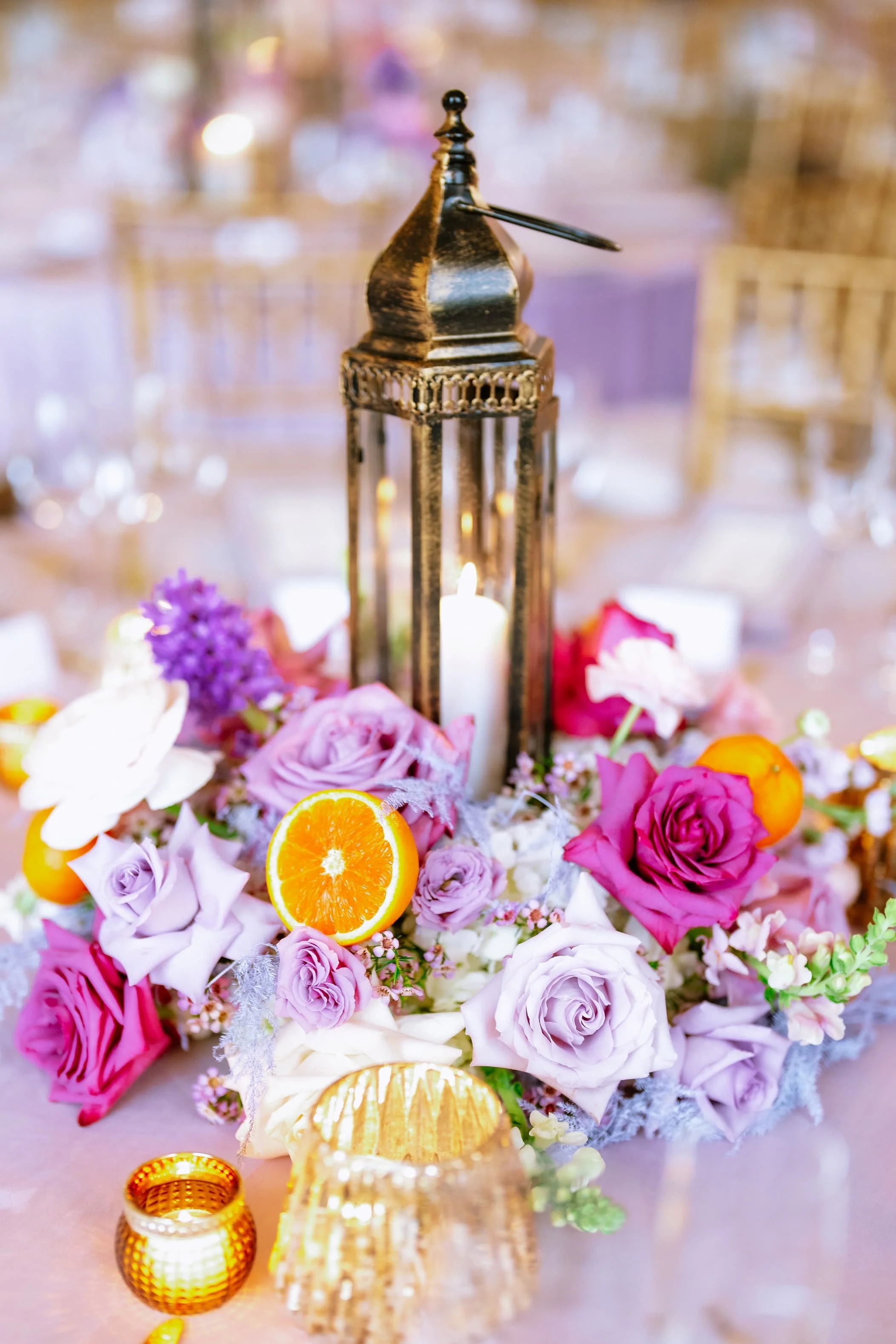 A decorative event table centerpiece with purple and pink roses, orange slices, candles, and a tall gold lantern at the center on a pink tablecloth.