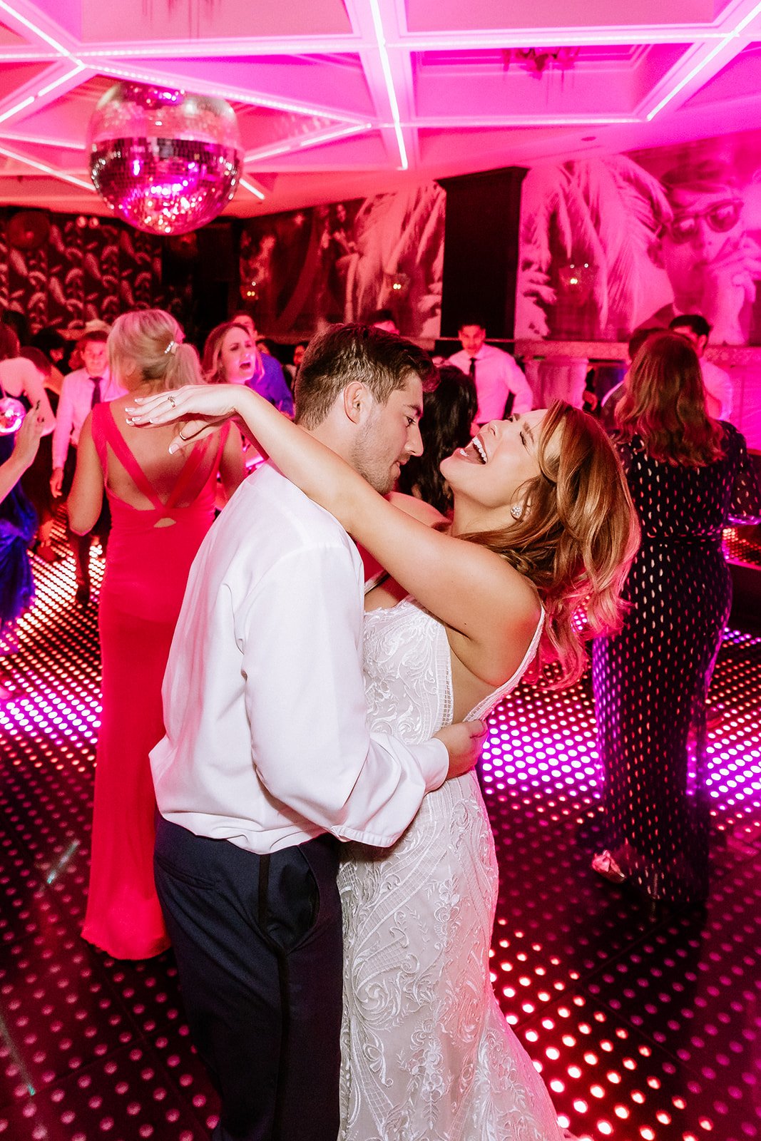 A couple dancing closely together at a lively party or wedding reception with colorful lights and other guests dancing in the background.