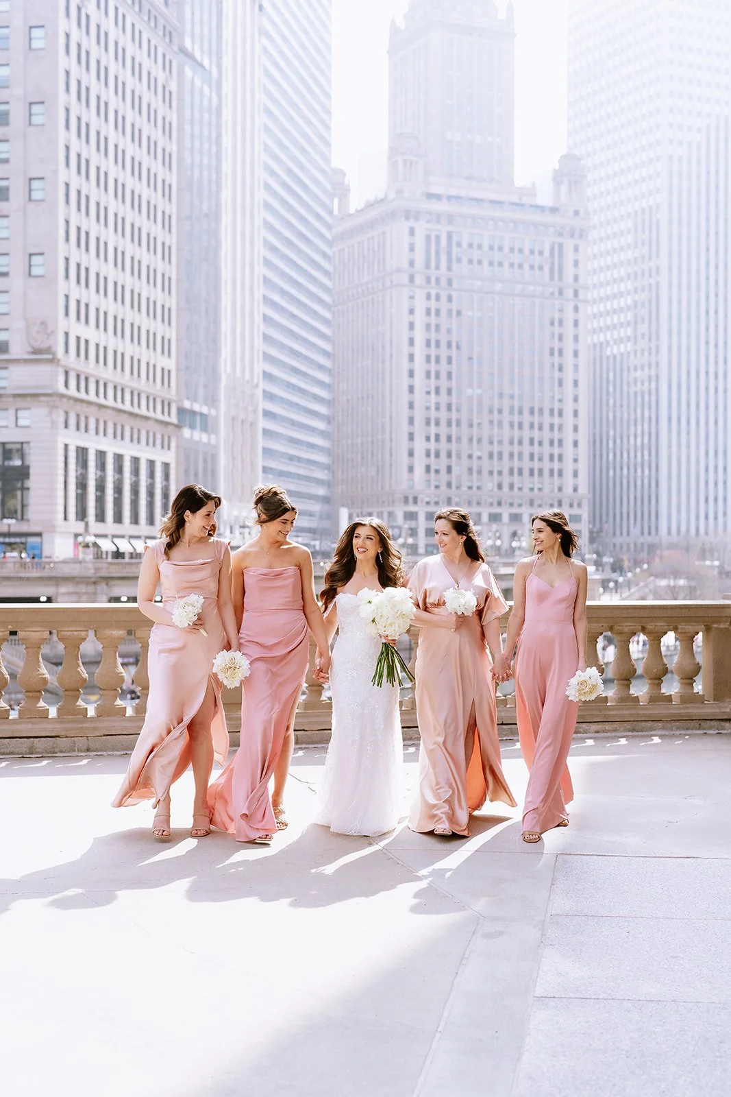 Bride in a white wedding dress walking with five bridesmaids in pink dresses on a city balcony with skyscrapers in the background