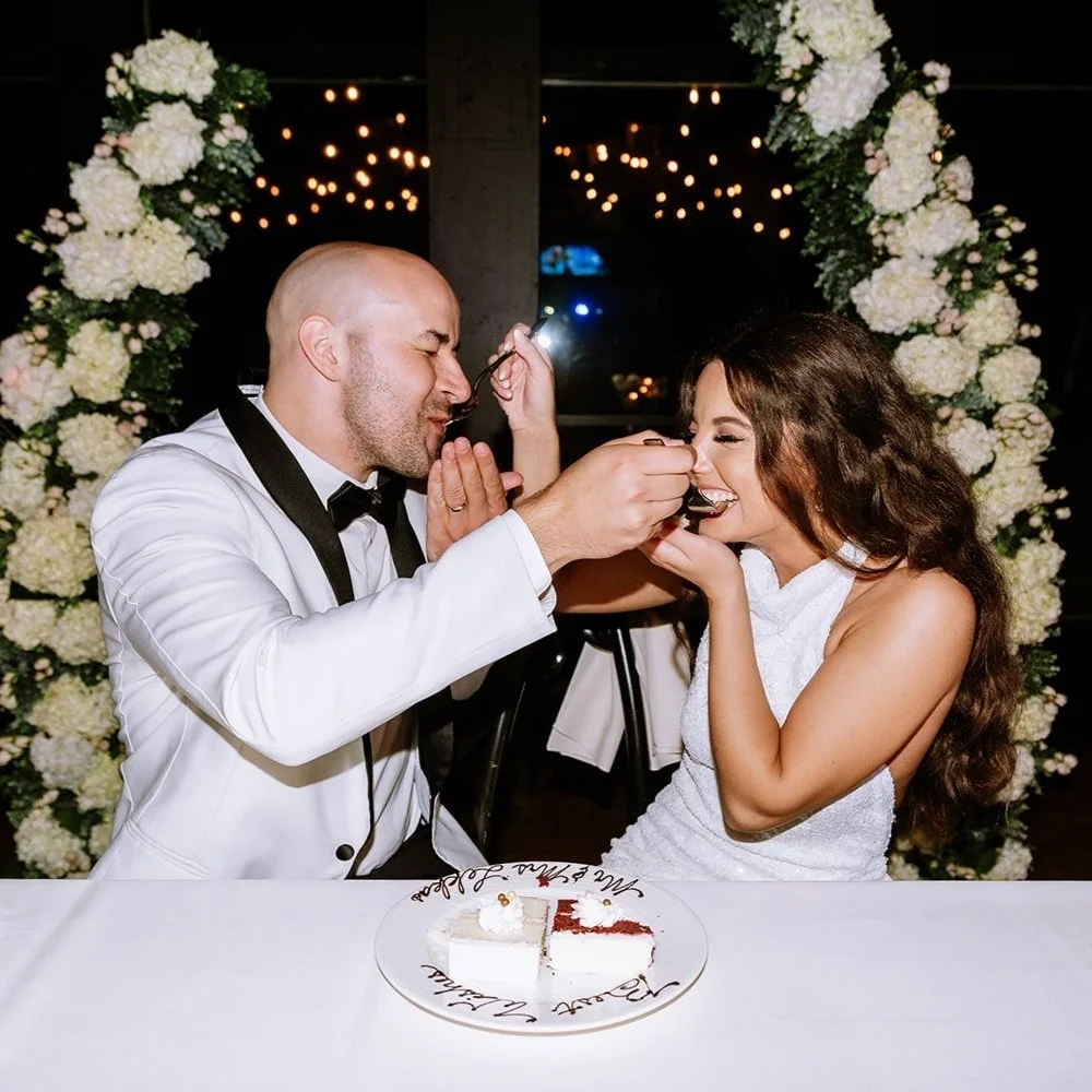 A man feeding a woman cake at a wedding celebration, with a floral arch and string lights in the background.