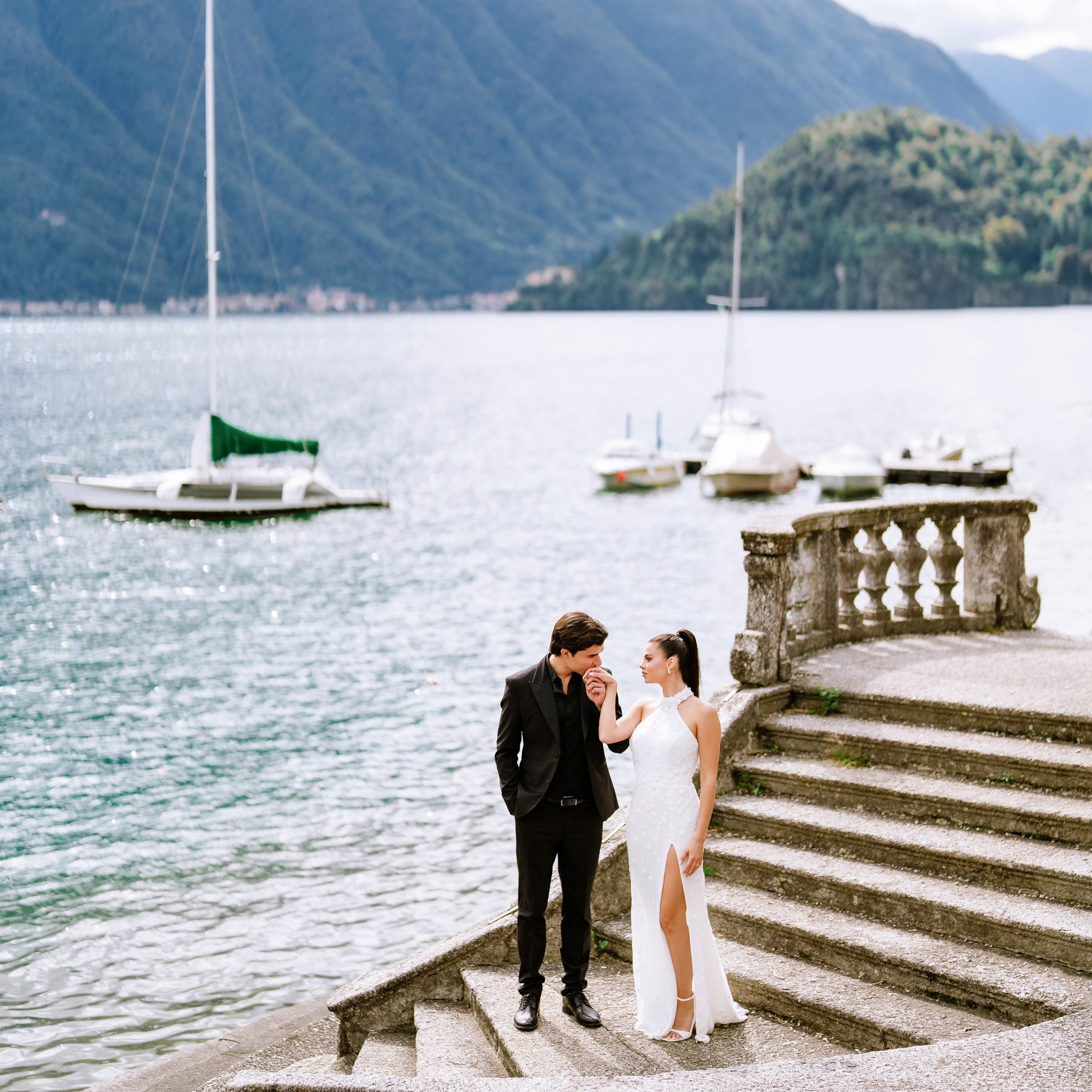 A couple dressed in formal attire on steps by a lake with sailboats and mountains in the background.
