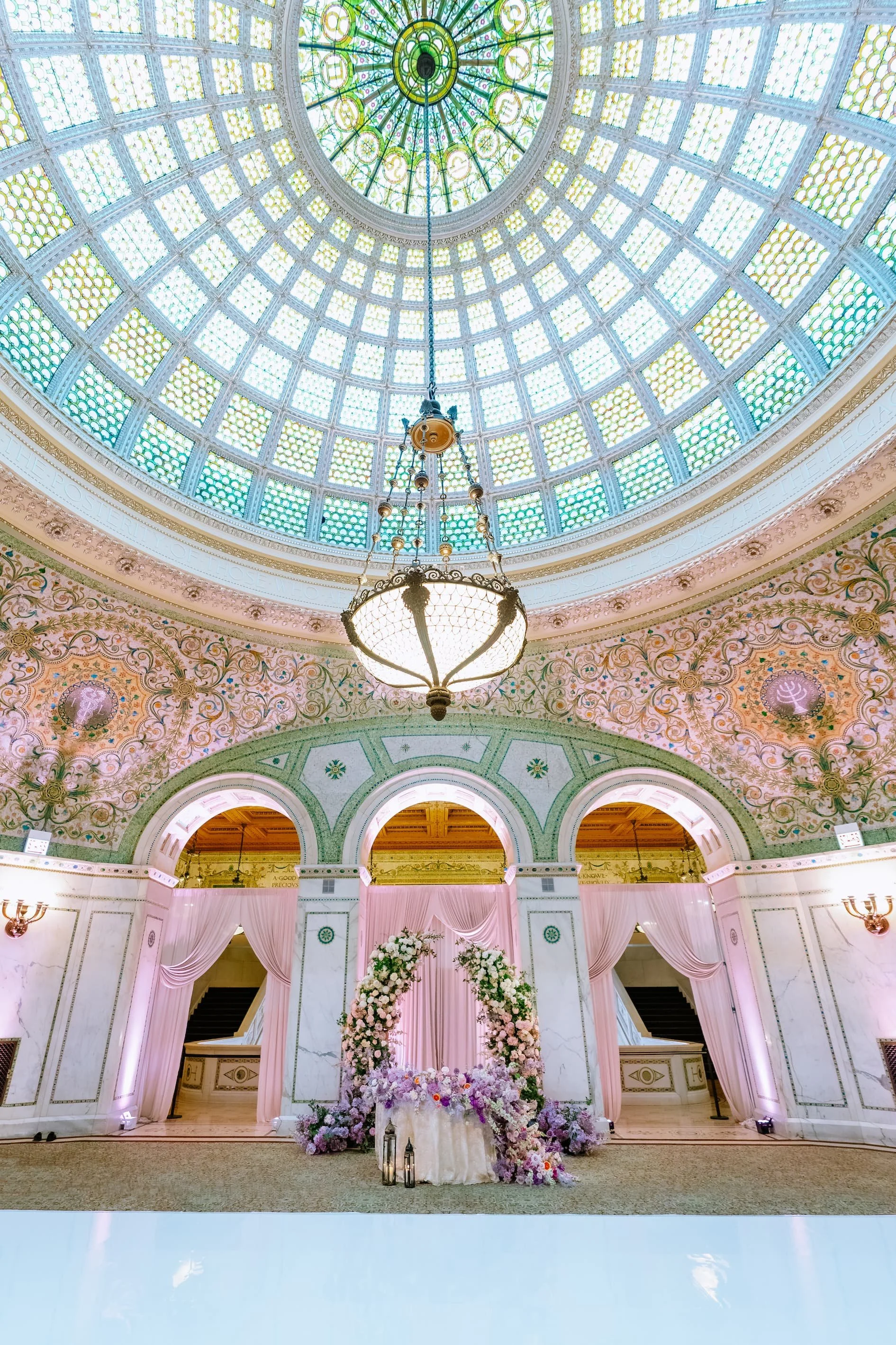 Elegant indoor event space with a high, stained glass dome ceiling, ornate walls decorated with floral and scroll patterns, pink curtains framing three archways, and a floral arch centerpiece with candles at a formal event or wedding.