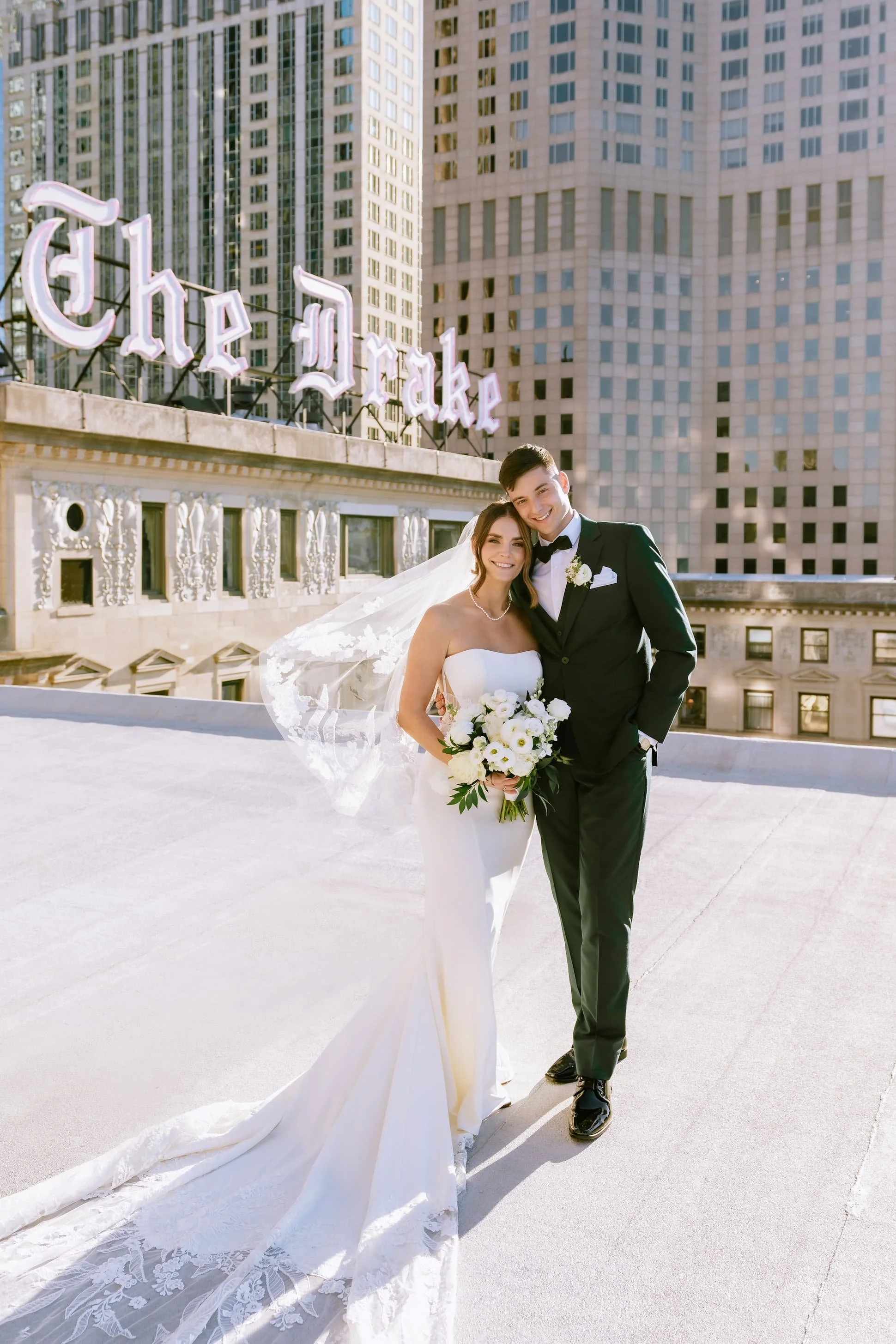 Bride and groom in wedding attire smiling on a rooftop with city skyscrapers and a neon sign reading 'The Drake' in the background.