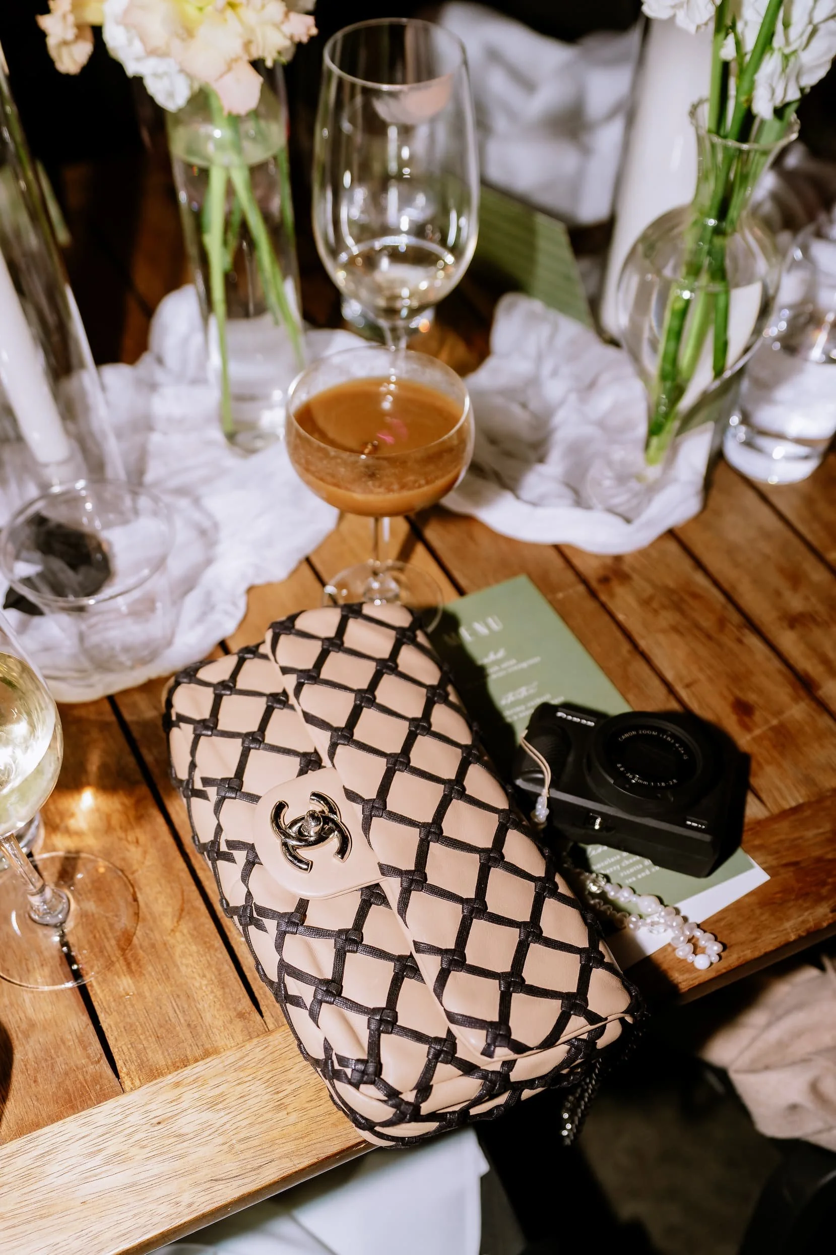 A wooden table set with various glasses, flowers in vases, a purse with a Chanel logo, a camera, and some printed material, possibly at a social gathering or event.