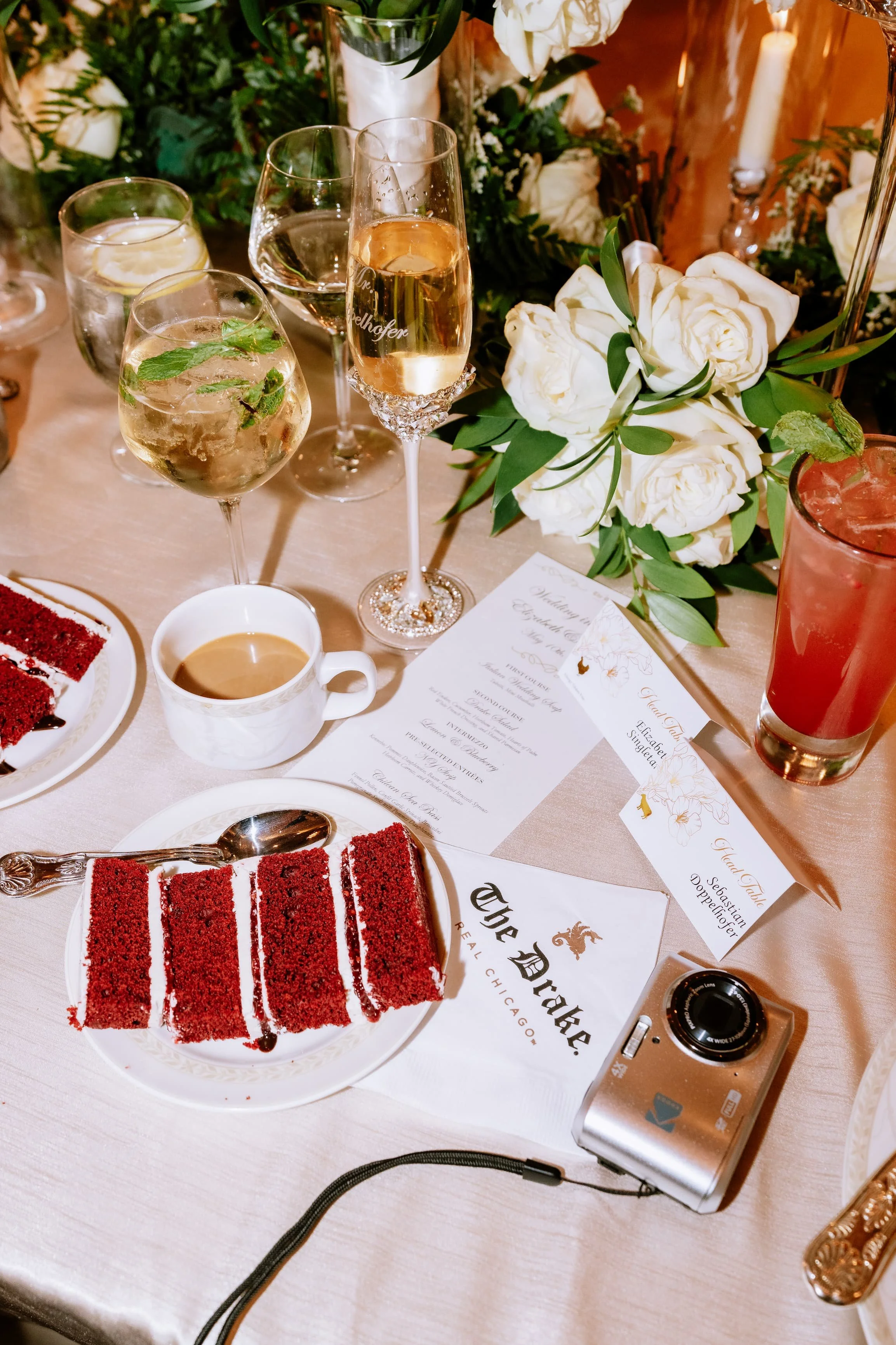 A table set for a celebration with various drinks, a slice of red velvet cake, wedding invitations, a camera, and floral arrangements with white roses and greenery.