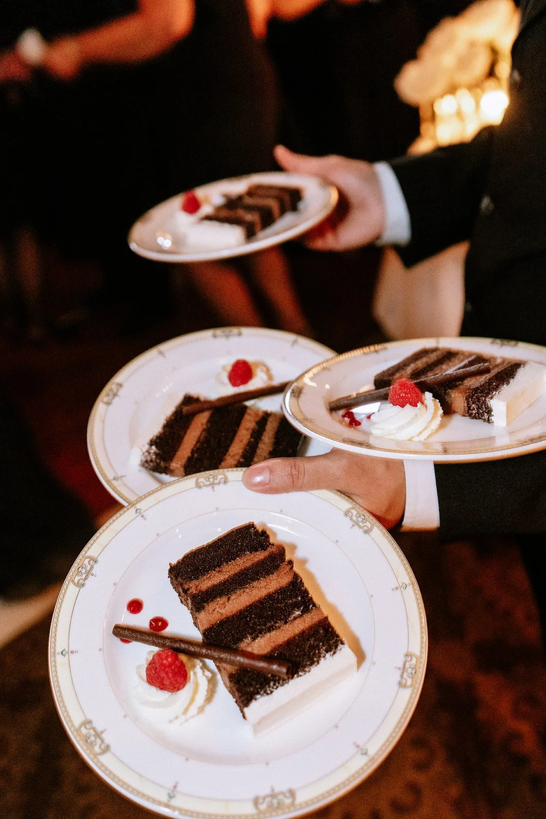 Multiple plates with slices of layered chocolate cake garnished with whipped cream, raspberries, and chocolate stick, being served at a formal event.