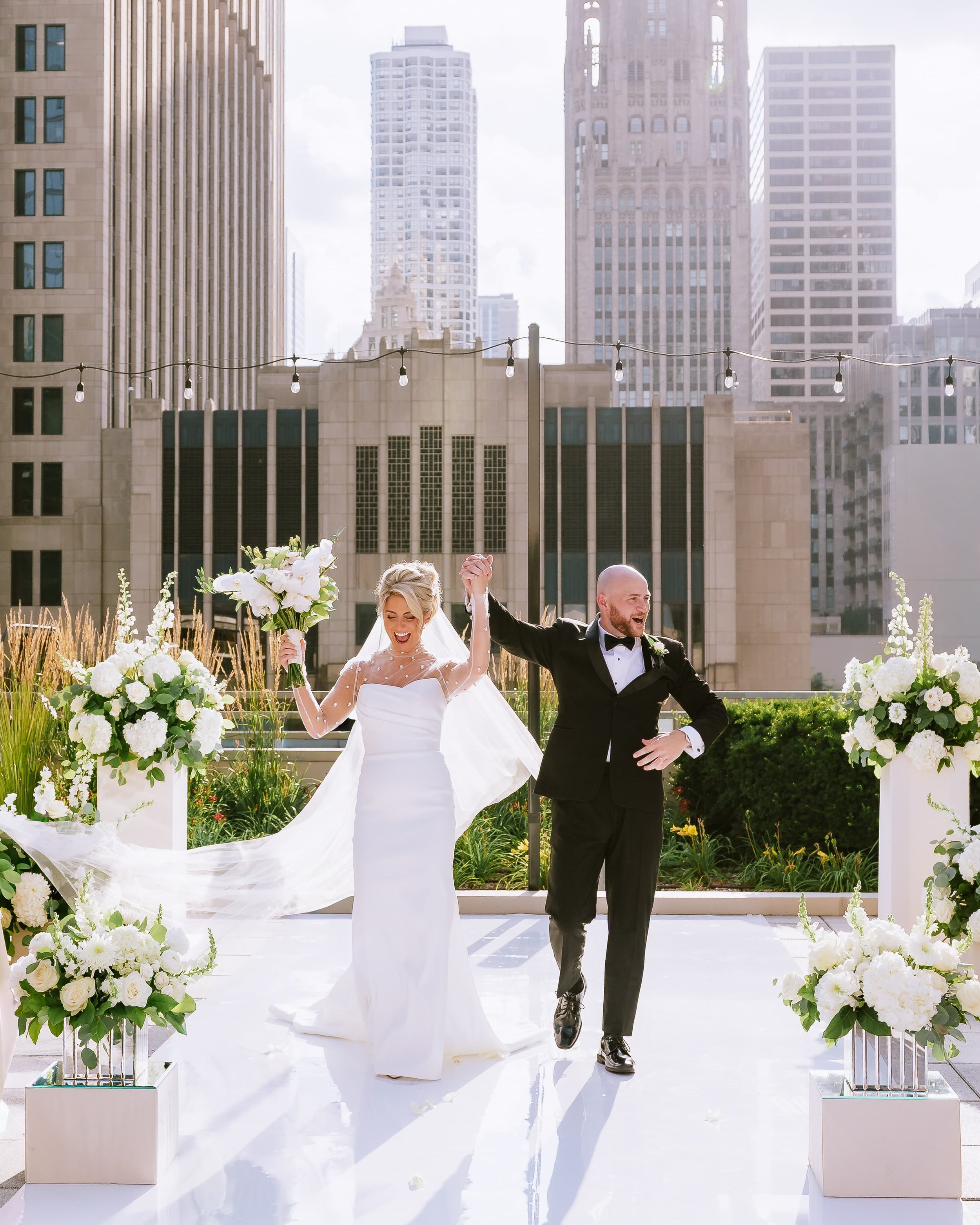 A newlywed couple in wedding attire walks down a rooftop aisle, holding hands and smiling, with city skyscrapers in the background and floral arrangements along the aisle.