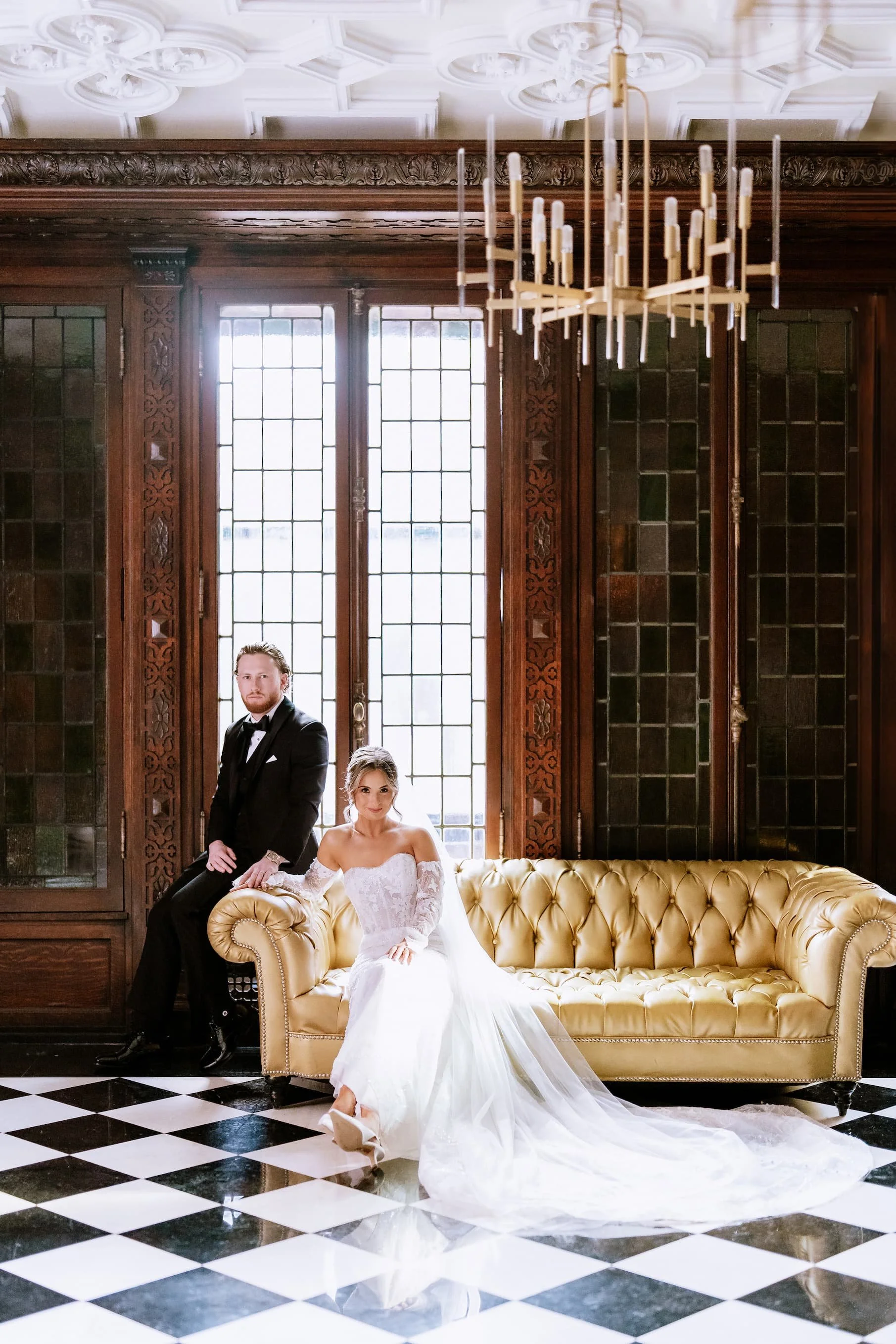 A bride in a white wedding gown sitting on a yellow tufted sofa and a groom in a black tuxedo leaning against the sofa in an ornate room with stained glass windows and a checkered black and white floor.