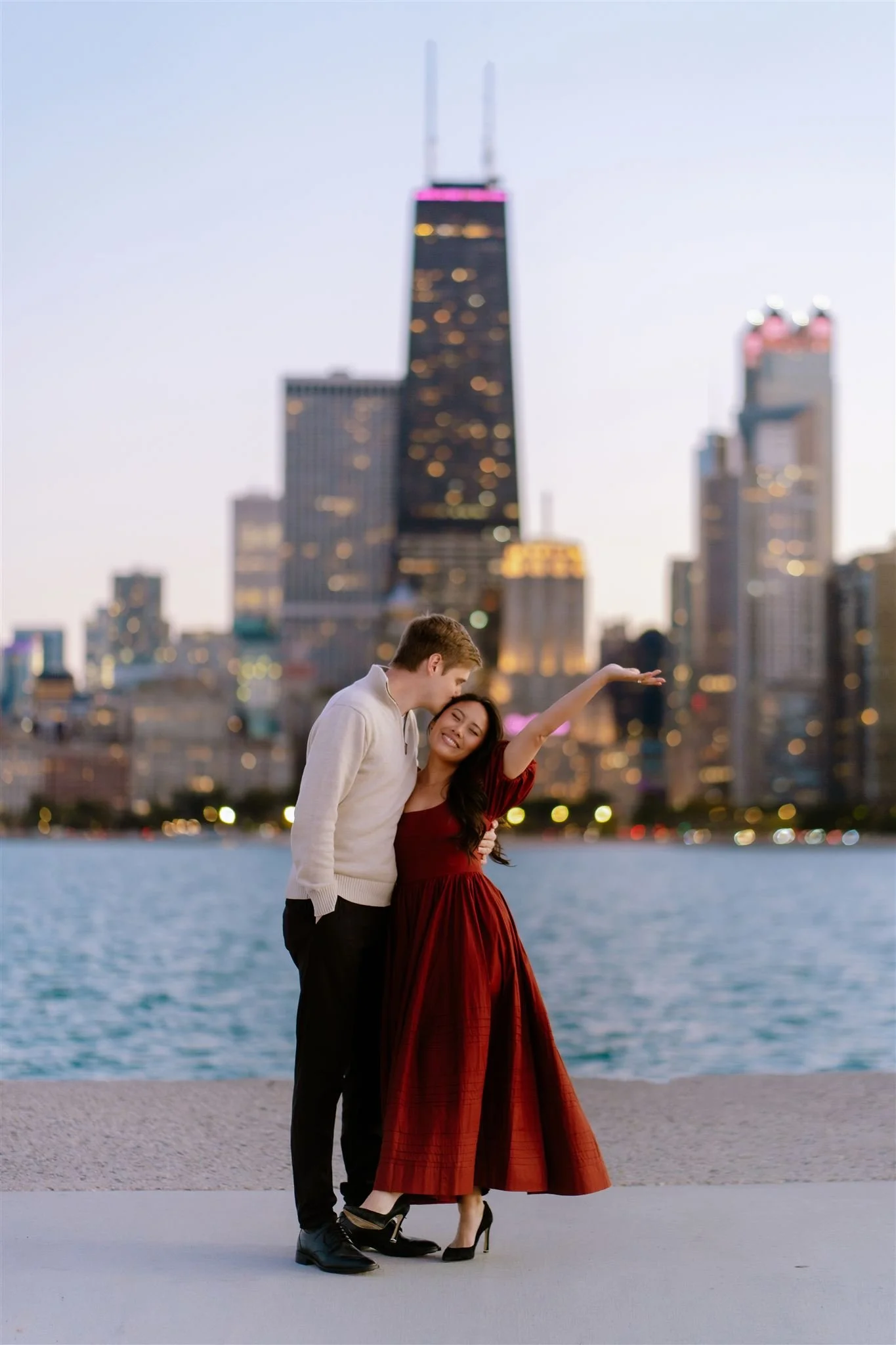 A young couple standing by a body of water in front of a city skyline during dusk; the woman is wearing a long red dress, and the man is dressed in a white sweater and dark pants.