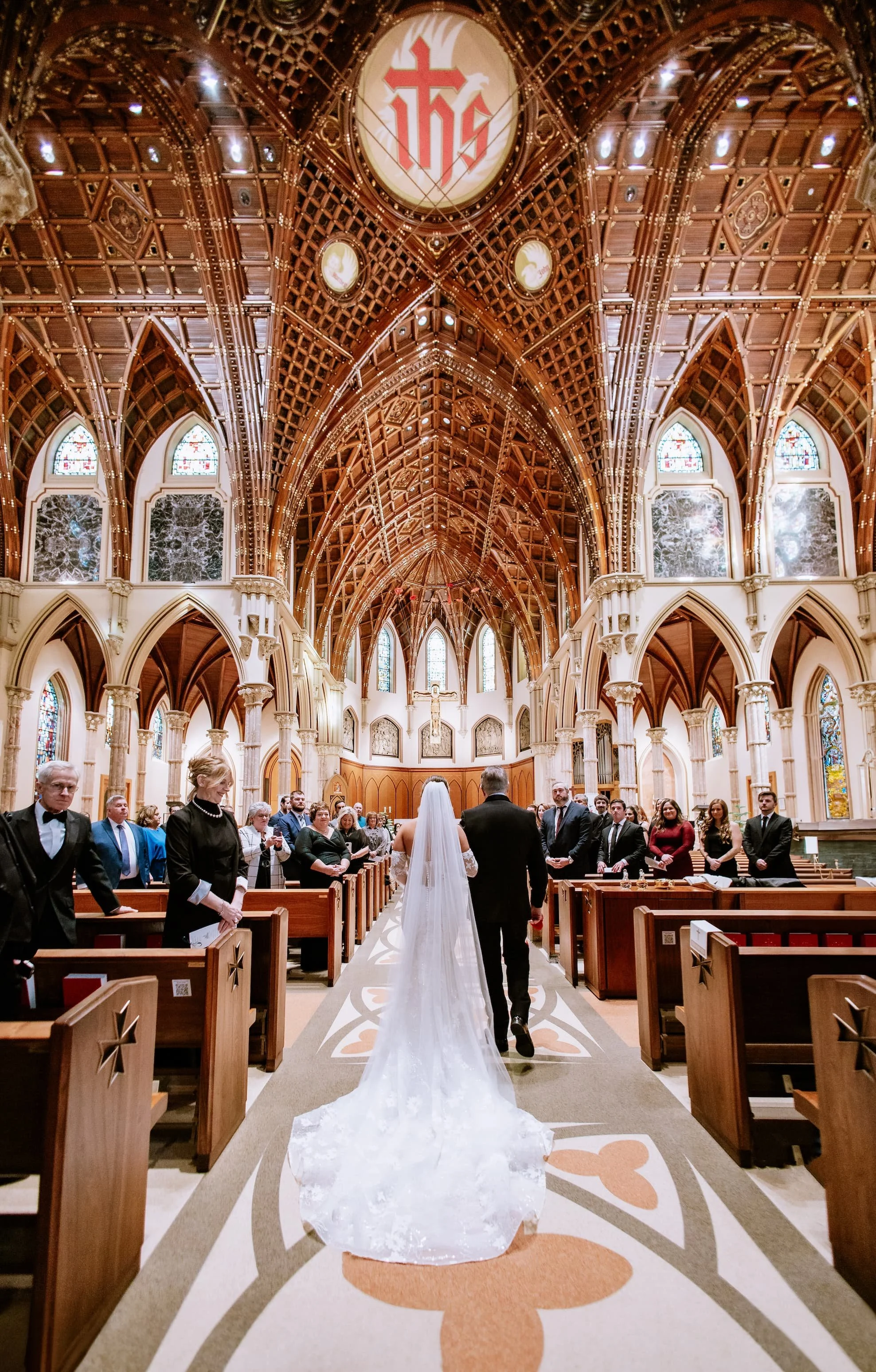 A bride and groom walking down the aisle of a church during their wedding ceremony. The church features a high, ornate wooden ceiling with stained glass windows and a large religious emblem. Wedding guests are standing on either side of the aisle, wa