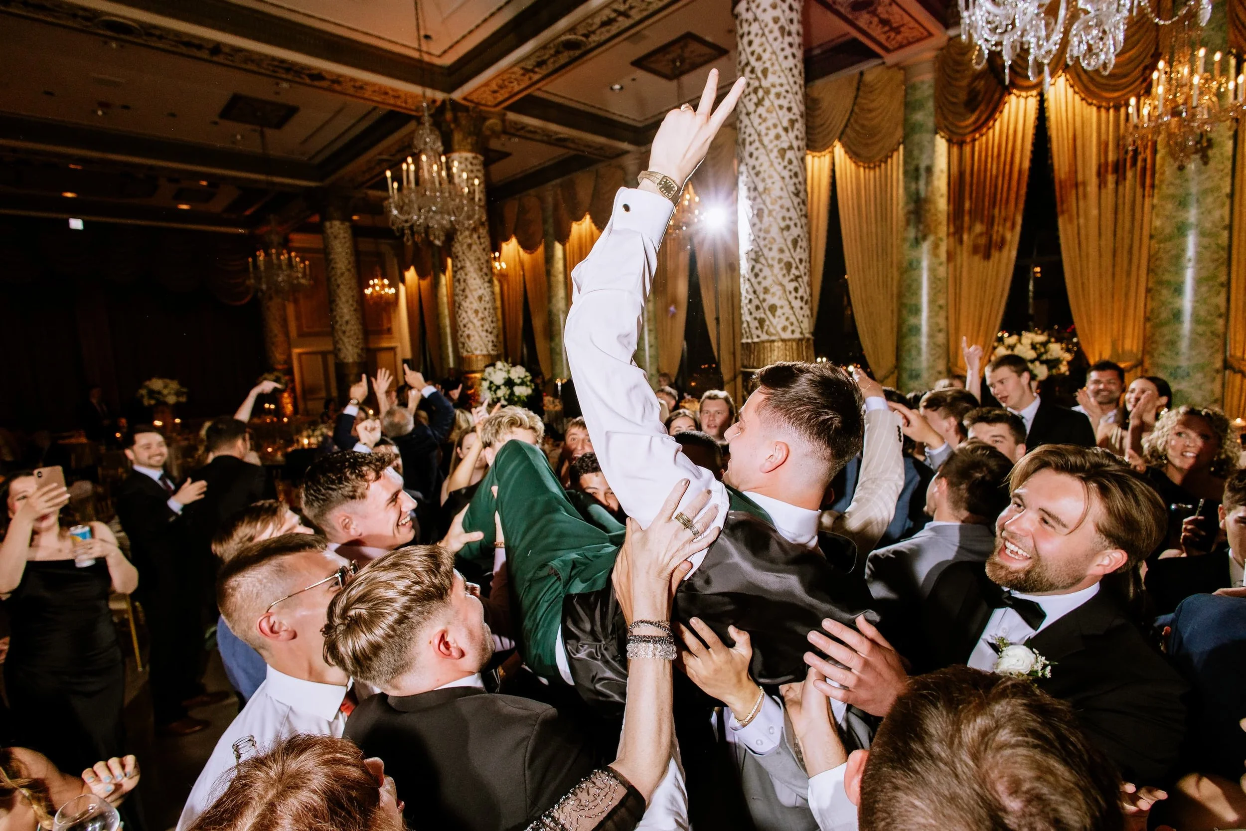 Celebration or party with a group of people, a man being lifted and cheering, in an elegant, decorated venue with chandeliers and curtains.